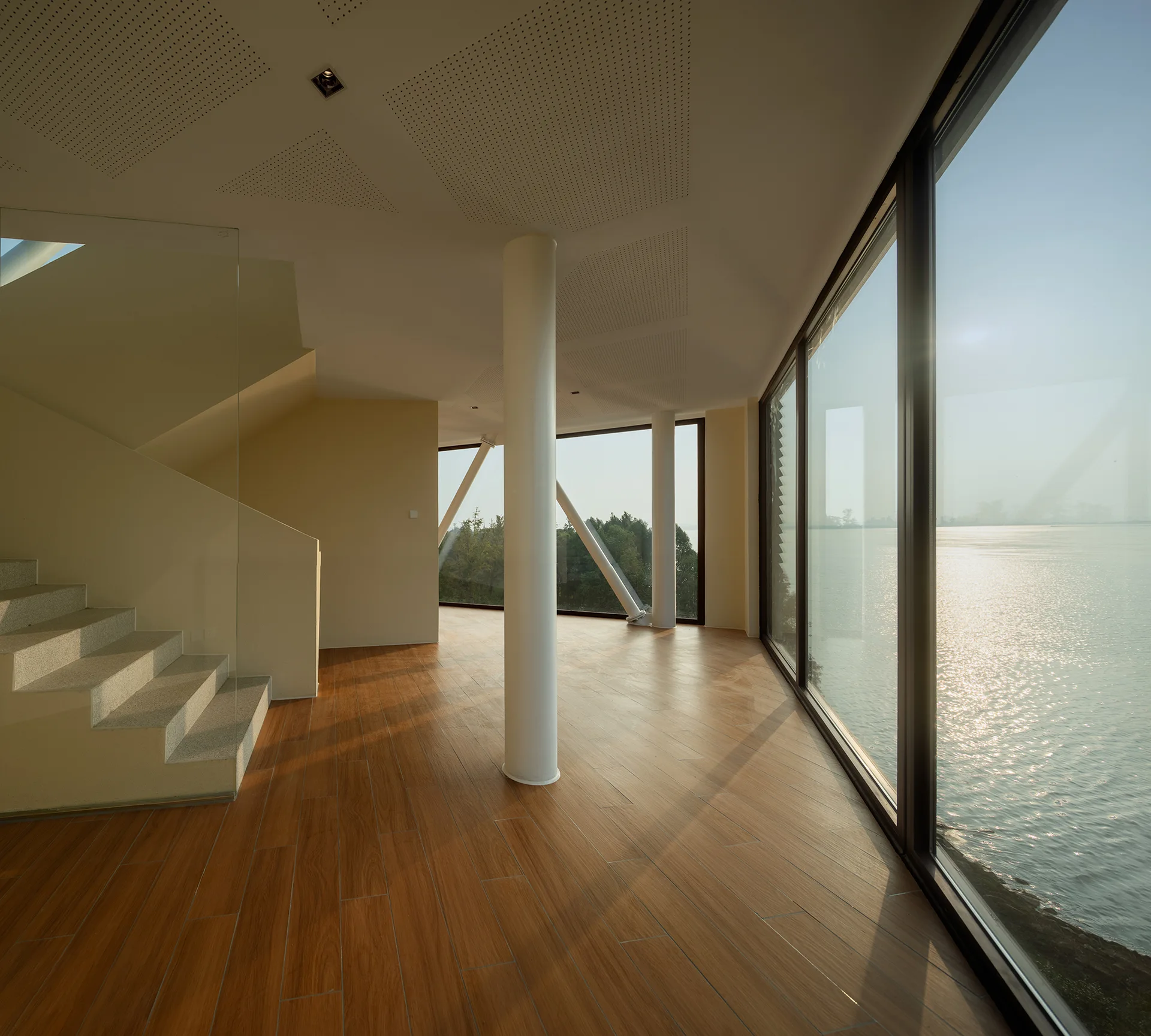 A modern minimalist interior with light wood flooring, a central structural column, a staircase, and massive glass windows looking out at water.