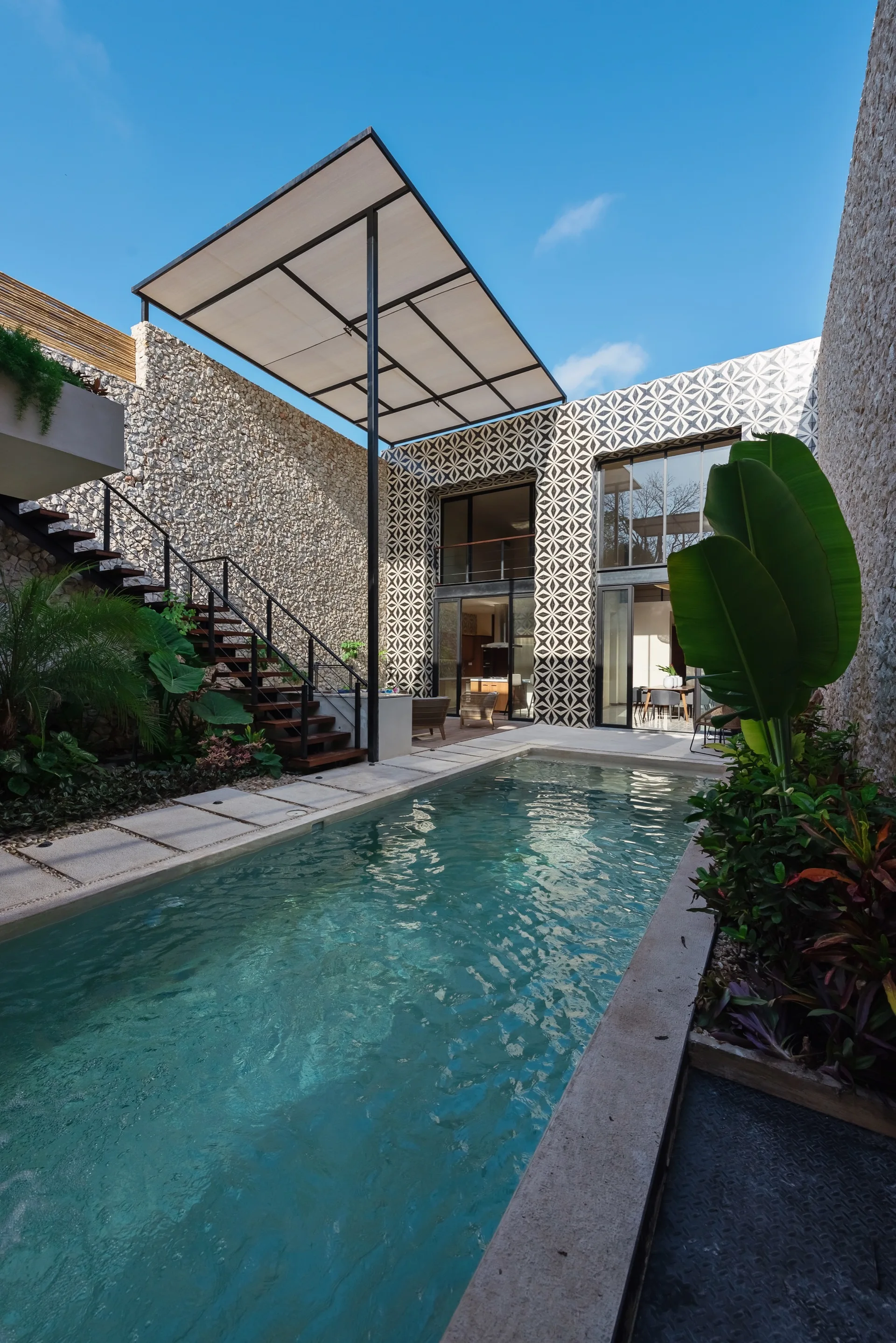 A courtyard swimming pool sits between rough stone walls and a black-and-white geometric tiled facade with a metal staircase.