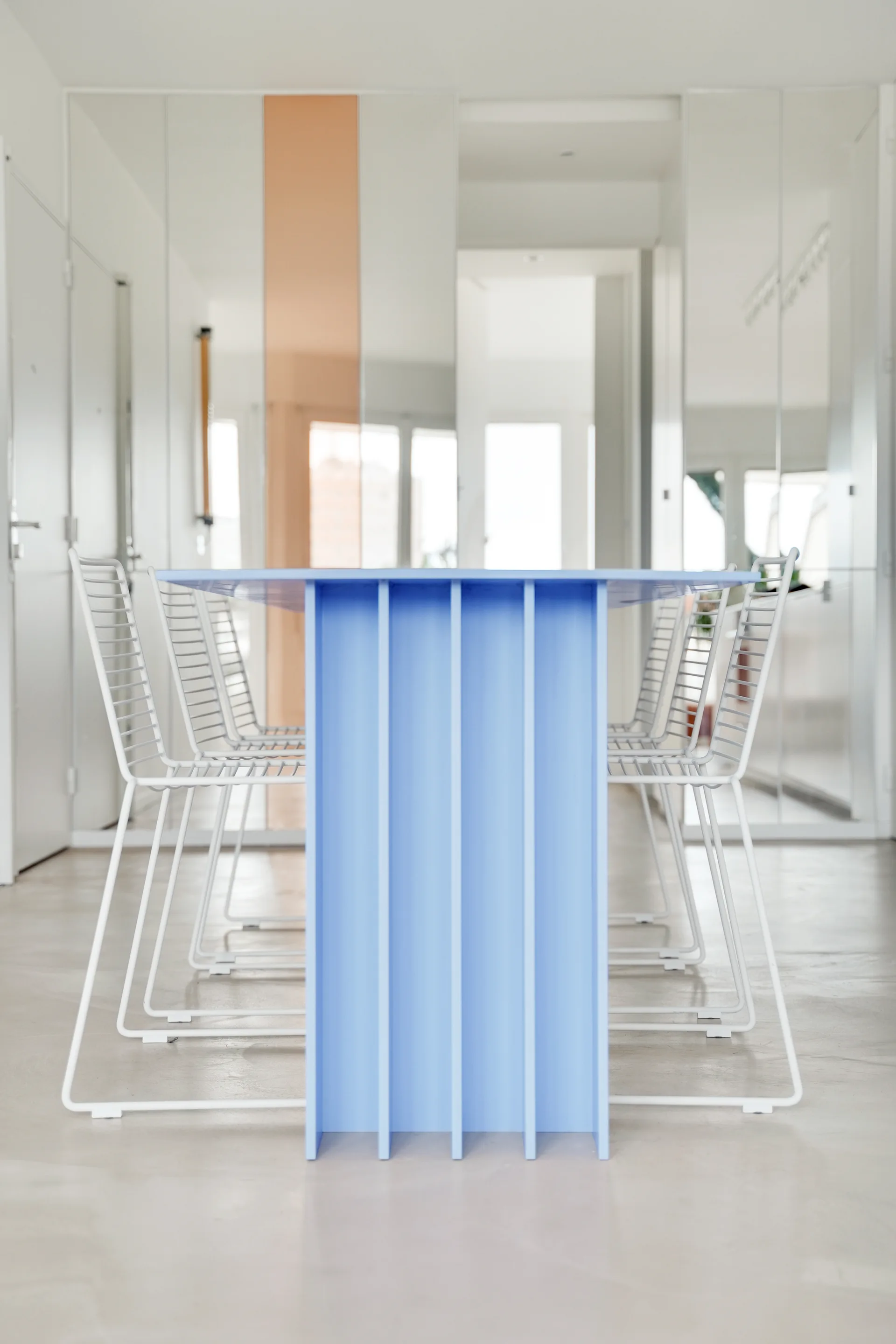 A minimalist dining table with a bright blue fin-style base is surrounded by white metal wire chairs in front of a mirrored wall.