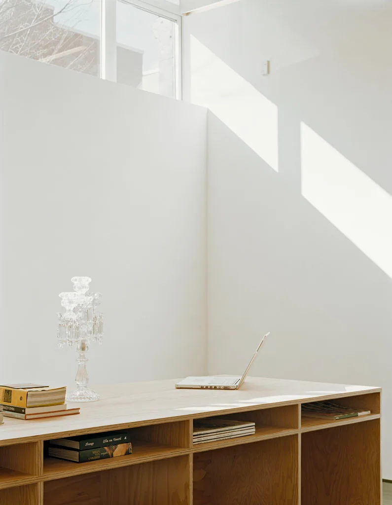 A light wood plywood desk with built-in storage cubbies holds a laptop and crystal candelabra under sunlight from an upper window.