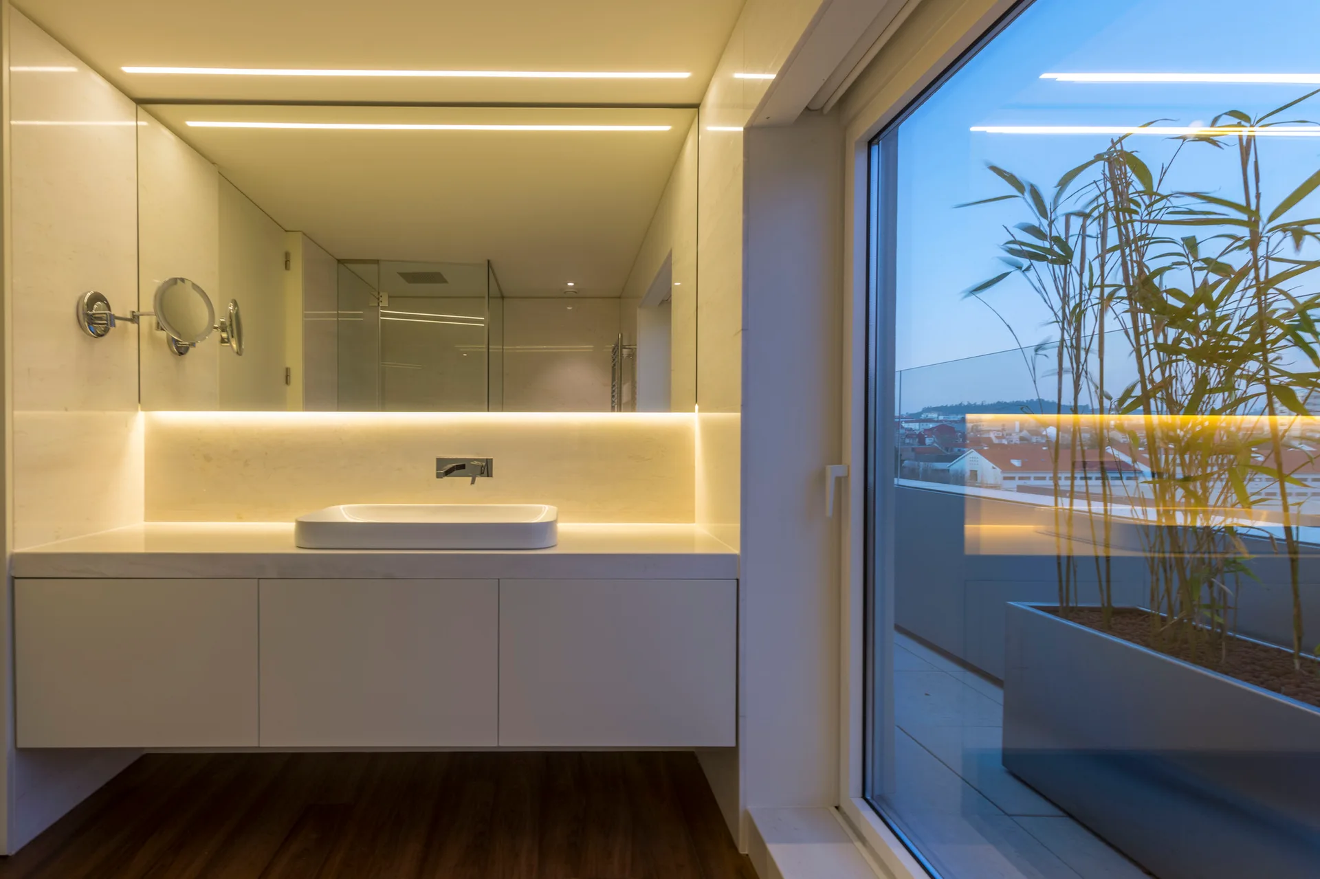 A minimalist white floating vanity with a backlit rectangular mirror stands next to a large sliding glass door revealing a balcony with potted bamboo.