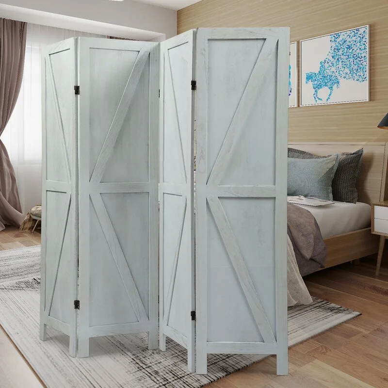 A white-washed wooden folding screen with an X-brace design stands on a textured rug in a bedroom with a light wood bed and blue pillows.