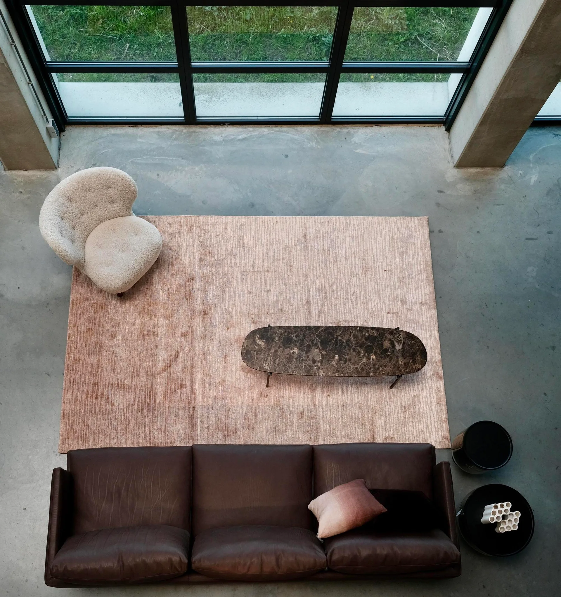 An overhead view of a living room with a brown leather sofa, a cream bouclé armchair, and an oval marble coffee table on a textured beige rug.