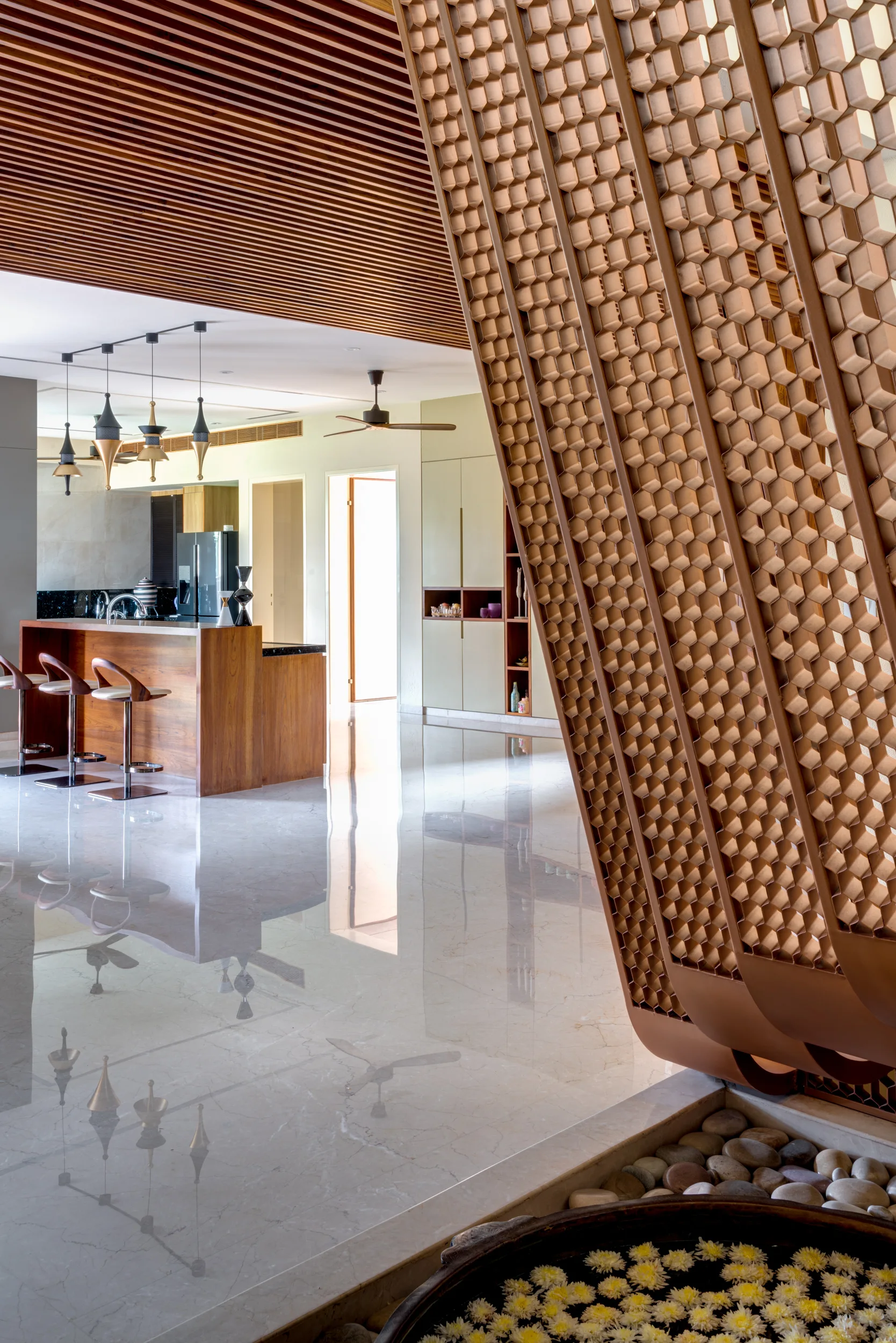 An open kitchen featuring a wooden island with bar stools, a highly reflective marble floor, a slatted wood ceiling, and a honeycomb-patterned screen.