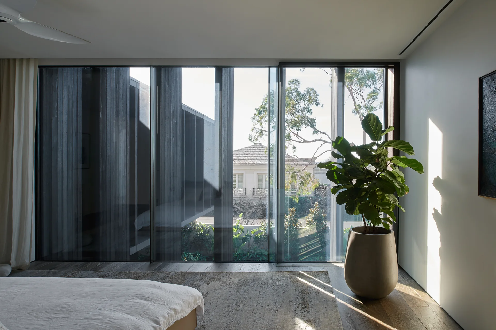 A minimalist bedroom features a bed, distressed rug, and a large potted fiddle-leaf fig plant next to floor-to-ceiling glass sliding doors.