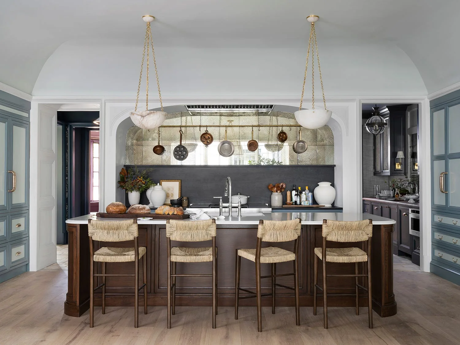 A dark wood kitchen island with four woven stools sits beneath pendant lights in front of an arched cooking niche with an antique mirror backsplash.