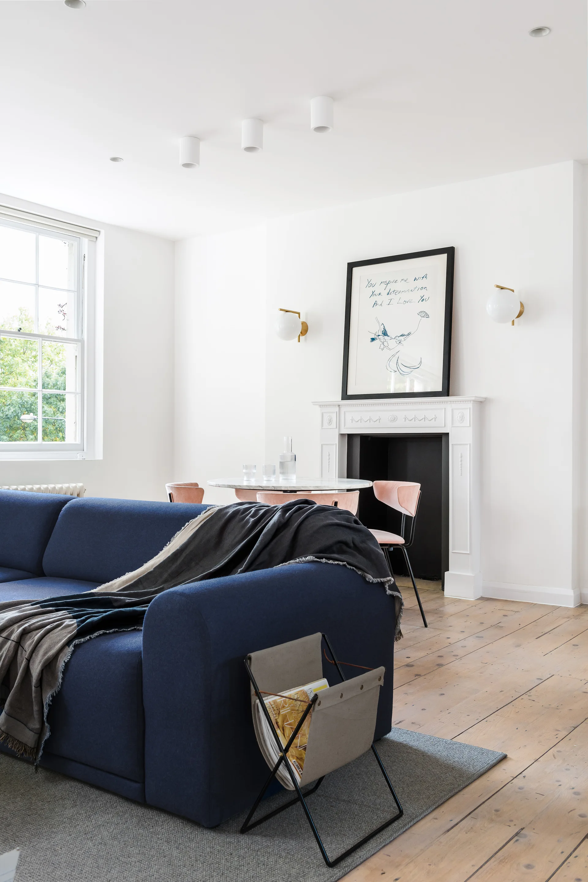 A bright living room features a blue sofa in the foreground and a white fireplace behind a small dining table with pink chairs.