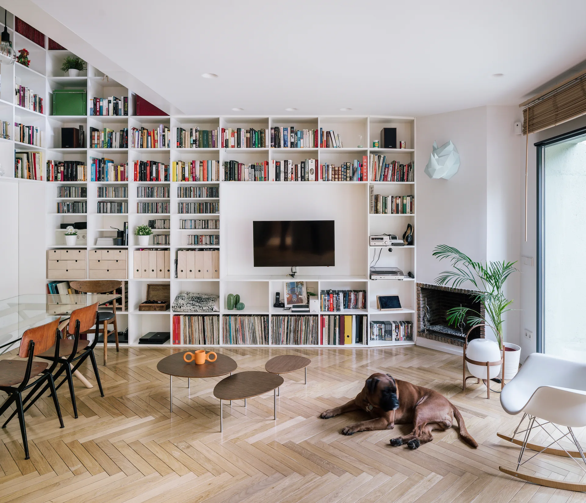 A bright living room features a massive wall-to-wall white bookshelf, herringbone pattern wood flooring, and a glass-topped dining table.