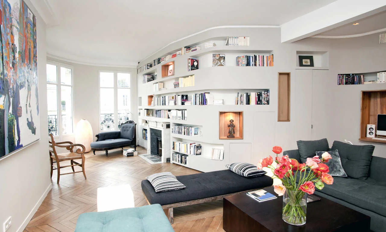 A grey living room with a curved built-in bookcase wrapping around the wall above a small fireplace, next to a black chaise lounge and dark sofas.