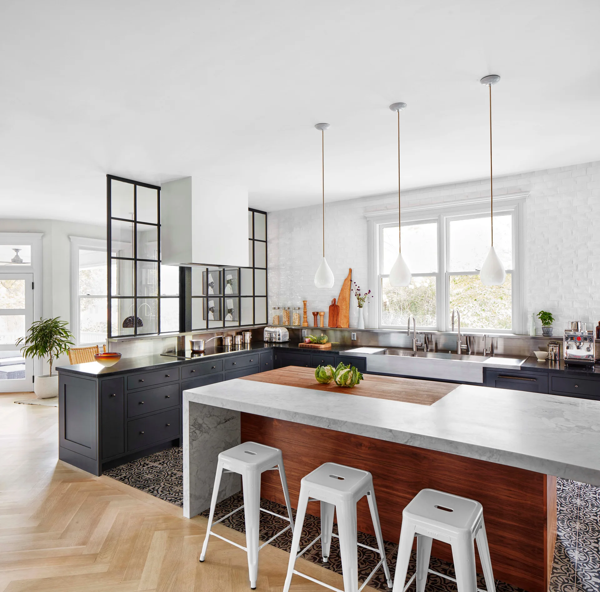 A modern kitchen featuring a marble and wood island with white stools, dark grey cabinetry, and a black-framed glass partition wall.