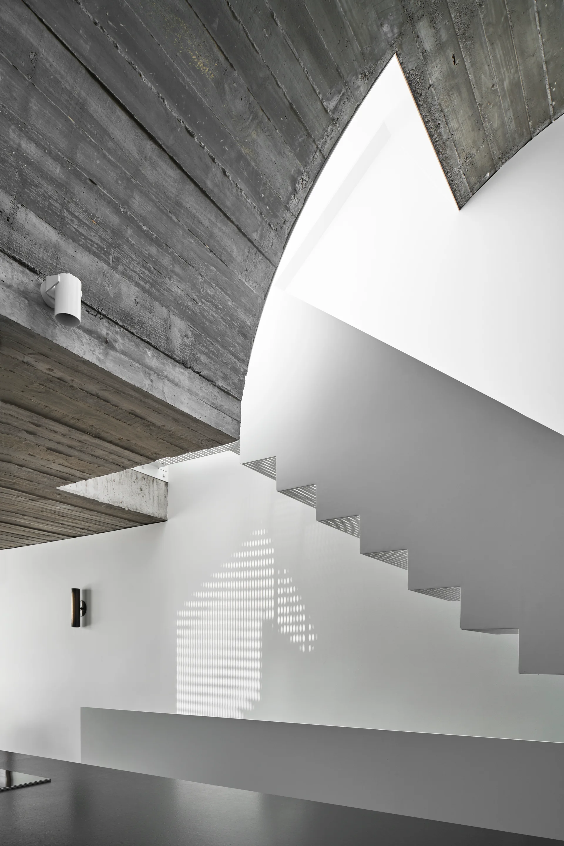 An angular white staircase ascends beneath a textured board-formed concrete ceiling, casting a dotted shadow onto a smooth white wall.