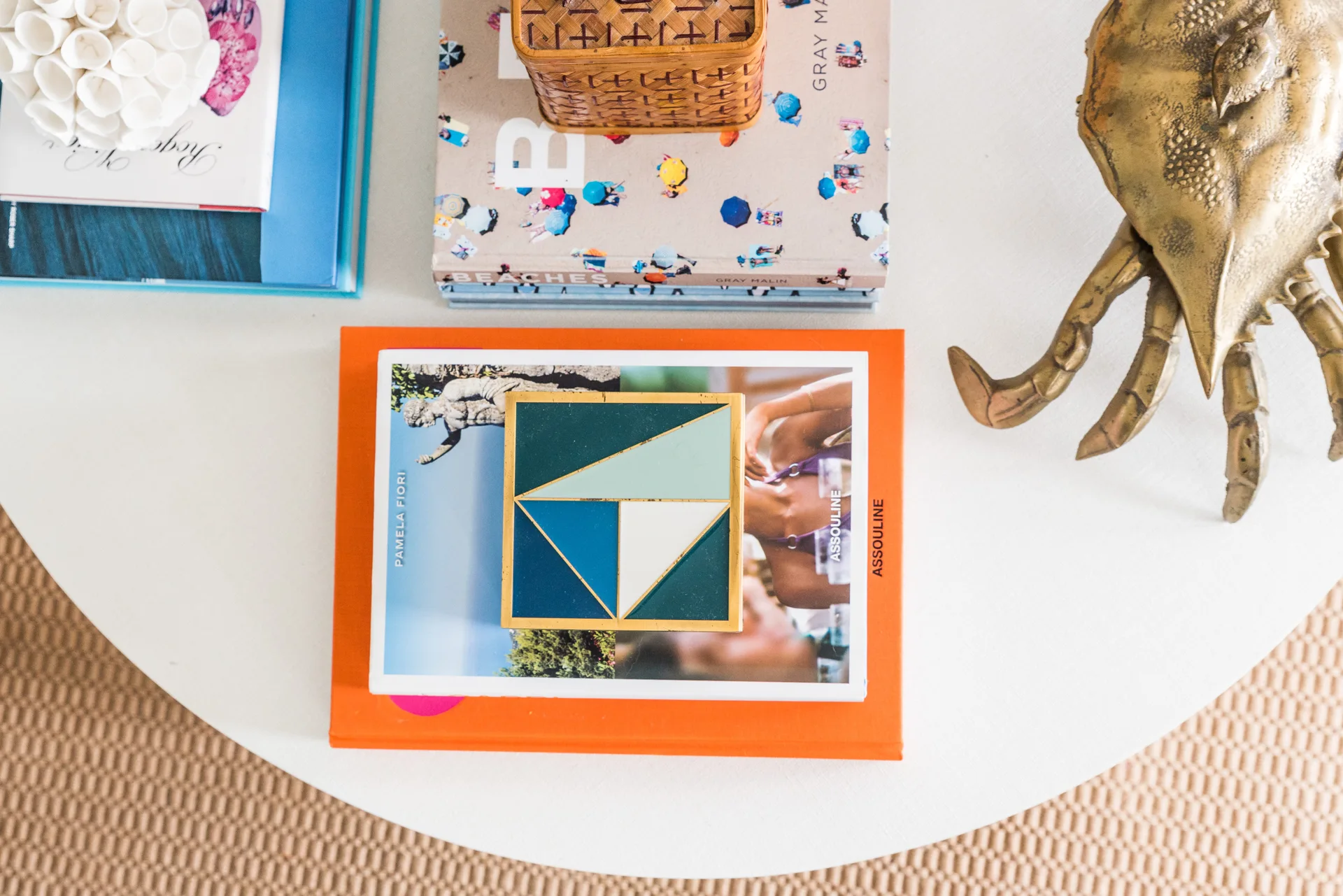 An overhead view of a round white coffee table styled with stacked art books, a brass crab sculpture, a wicker box, and white coral.