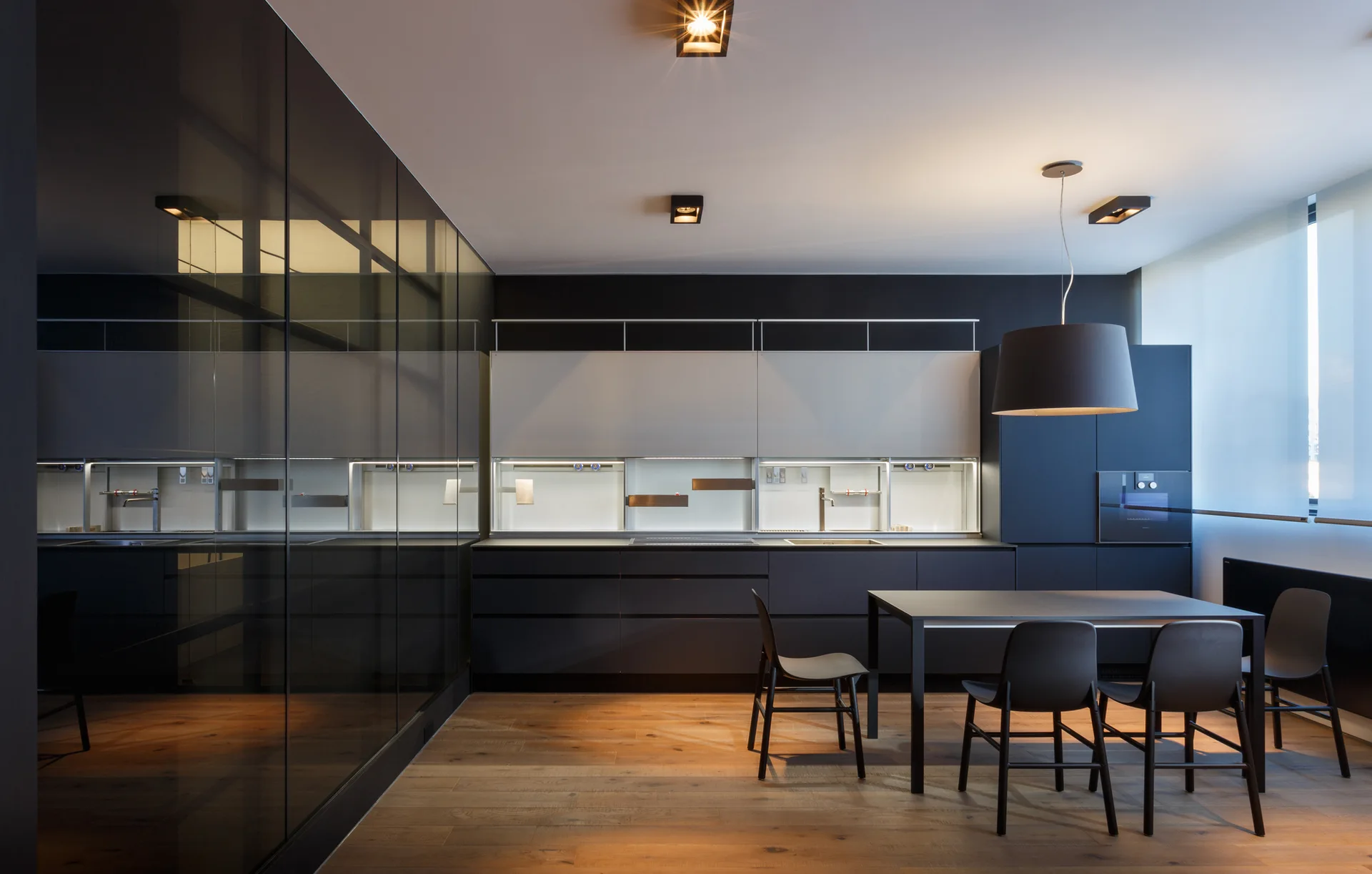 A minimalist dark gray kitchen features integrated cabinetry, a large reflective mirrored wall, a black dining set, and light wood plank flooring.