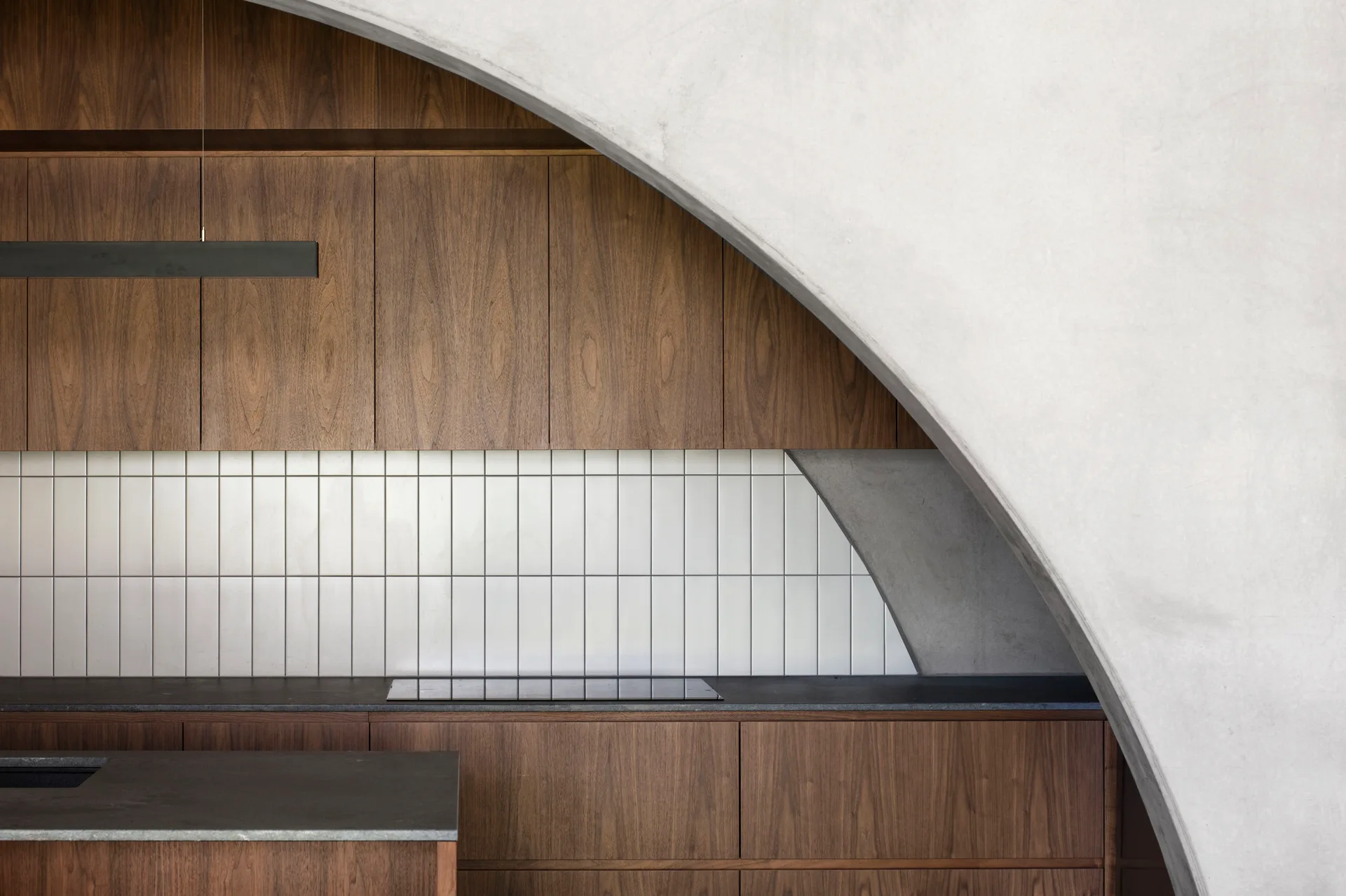 A modern kitchen featuring warm wood cabinetry, a dark countertop, a vertical white tile backsplash, and a prominent grey concrete arch.