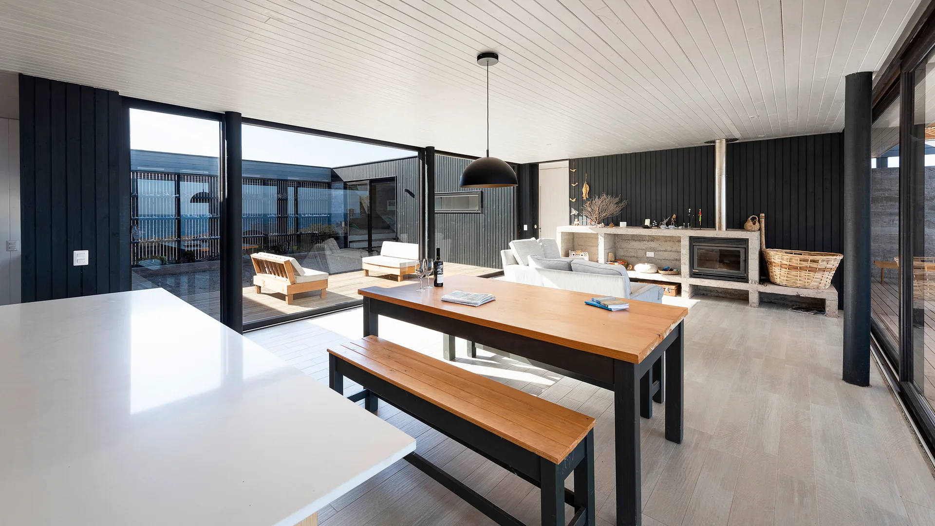 A modern open-plan living and dining room features black slatted walls, a concrete fireplace, a wooden dining table, and large sliding glass doors.