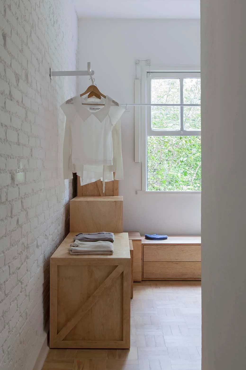 A minimalist closet features a white painted brick wall, a wall-mounted clothing rack, and light wood storage crates beneath a window.