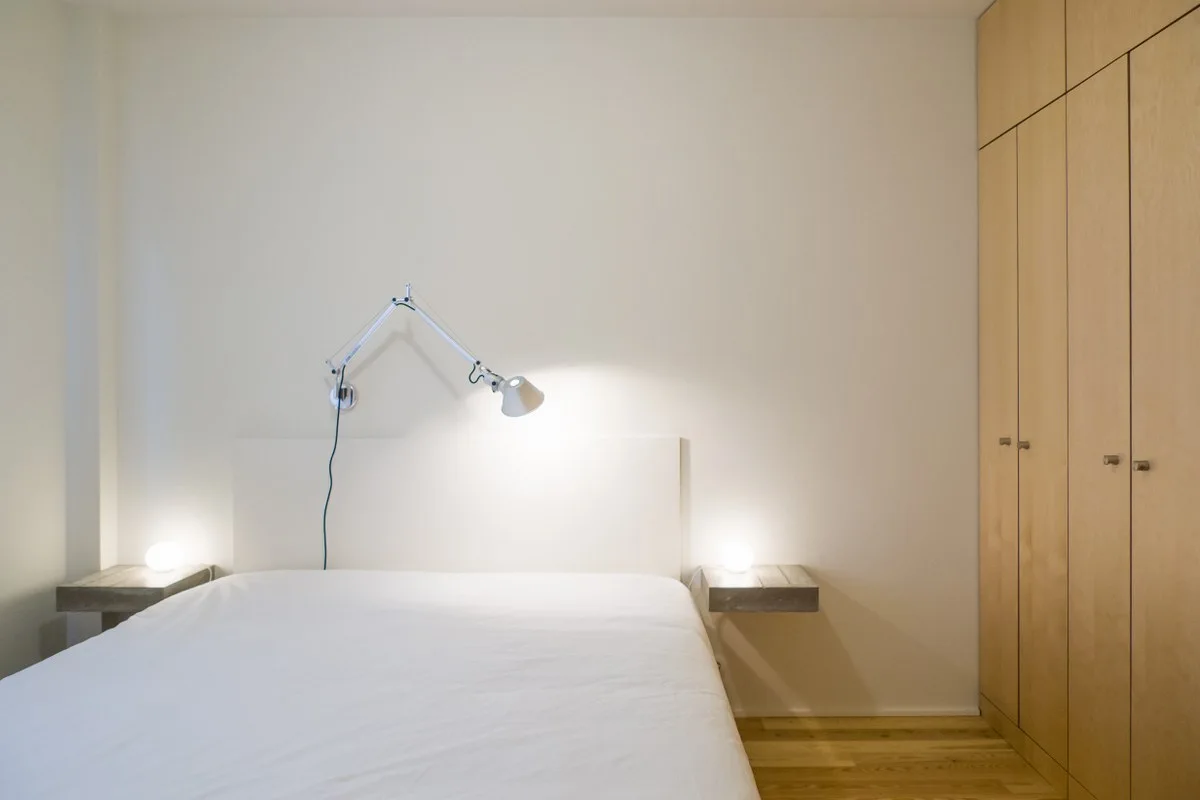 A minimalist bedroom featuring a bed with wooden floating nightstands, small glowing spherical lamps, and a tall built-in wood wardrobe.