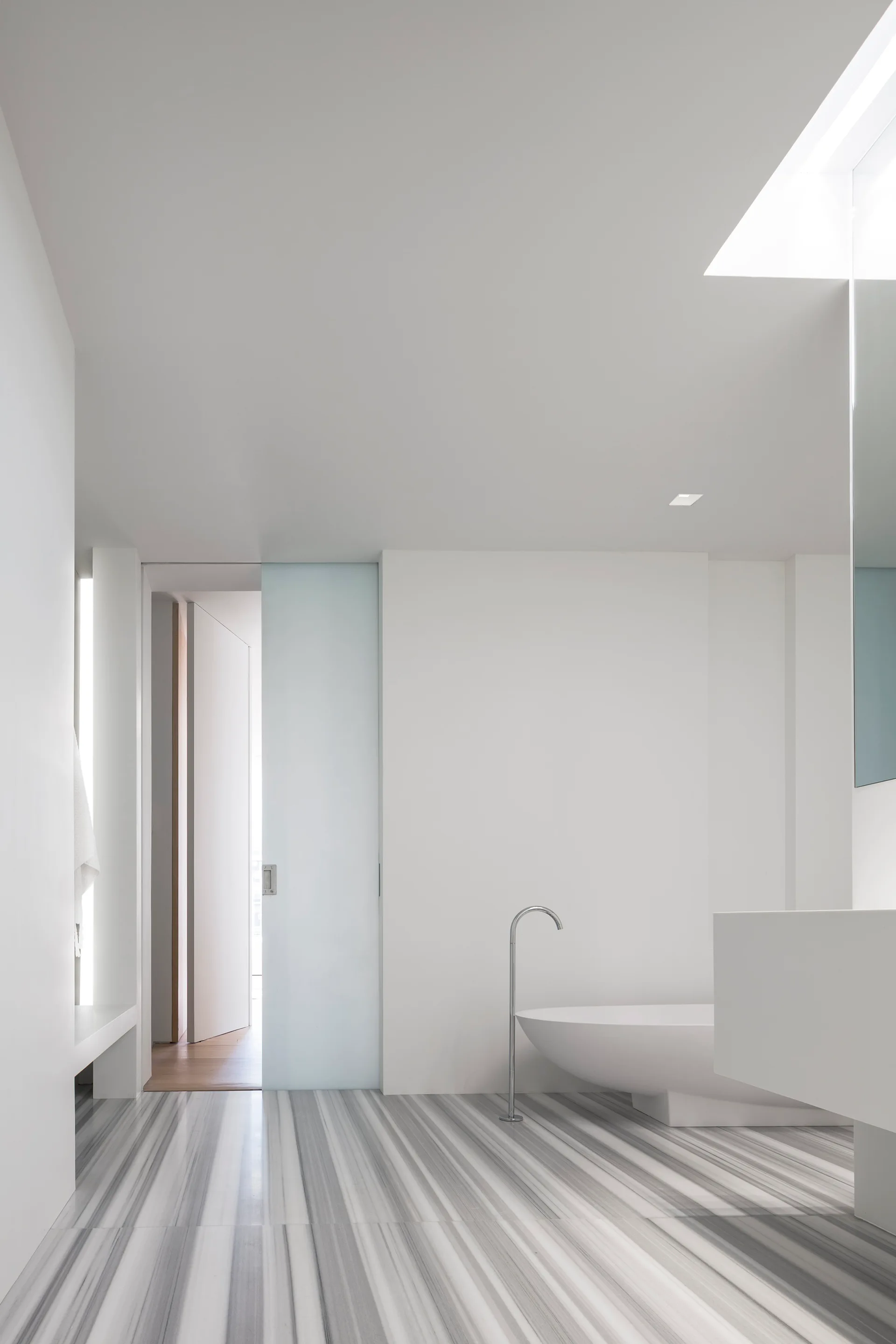A minimalist white bathroom features a freestanding bathtub on a linear striped marble floor next to a sliding frosted glass door.