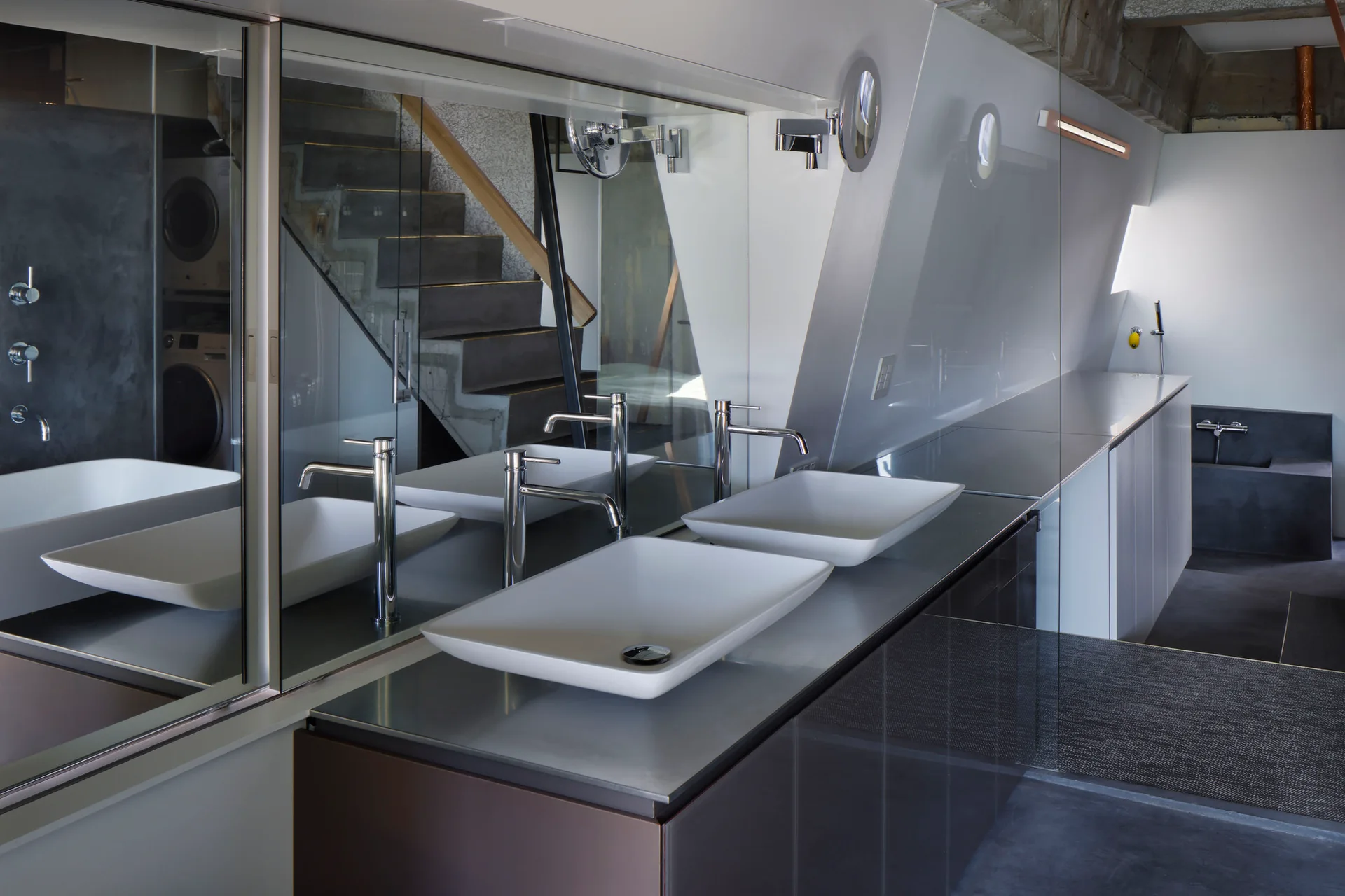 A minimalist bathroom vanity features two white rectangular vessel sinks on a metal countertop beneath a large mirror reflecting a staircase.