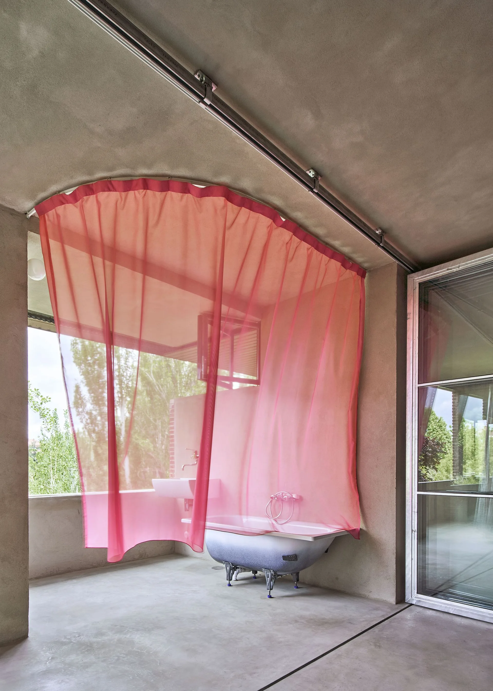 A minimalist grey concrete bathroom with a clawfoot bathtub partially screened by a sheer pink curtain hanging from a curved ceiling track.