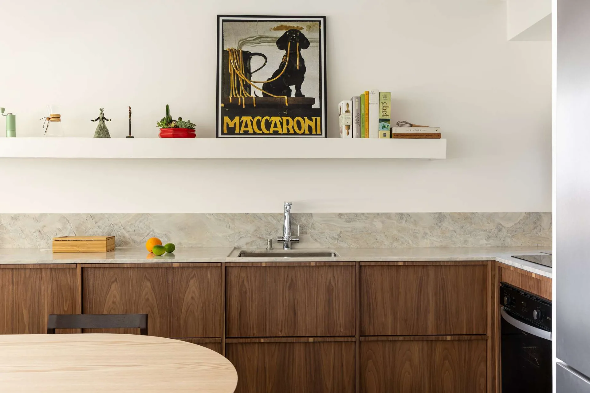 A modern kitchen features walnut wood lower cabinets, a light grey veined stone backsplash, and a long white floating shelf displaying a vintage poster.