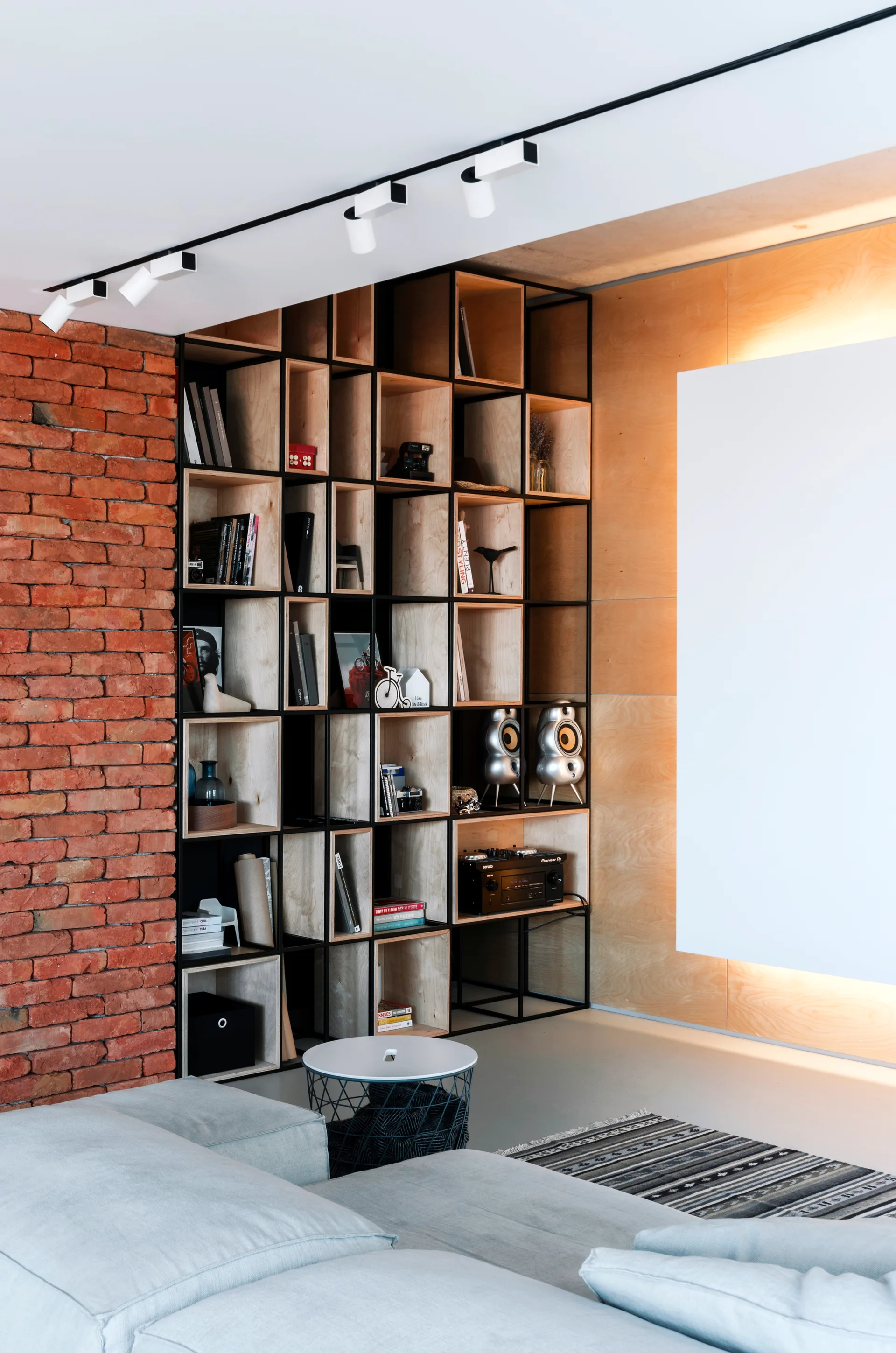 A modern living room features a tall black metal and plywood shelving unit set against an exposed red brick wall and light wood paneling.