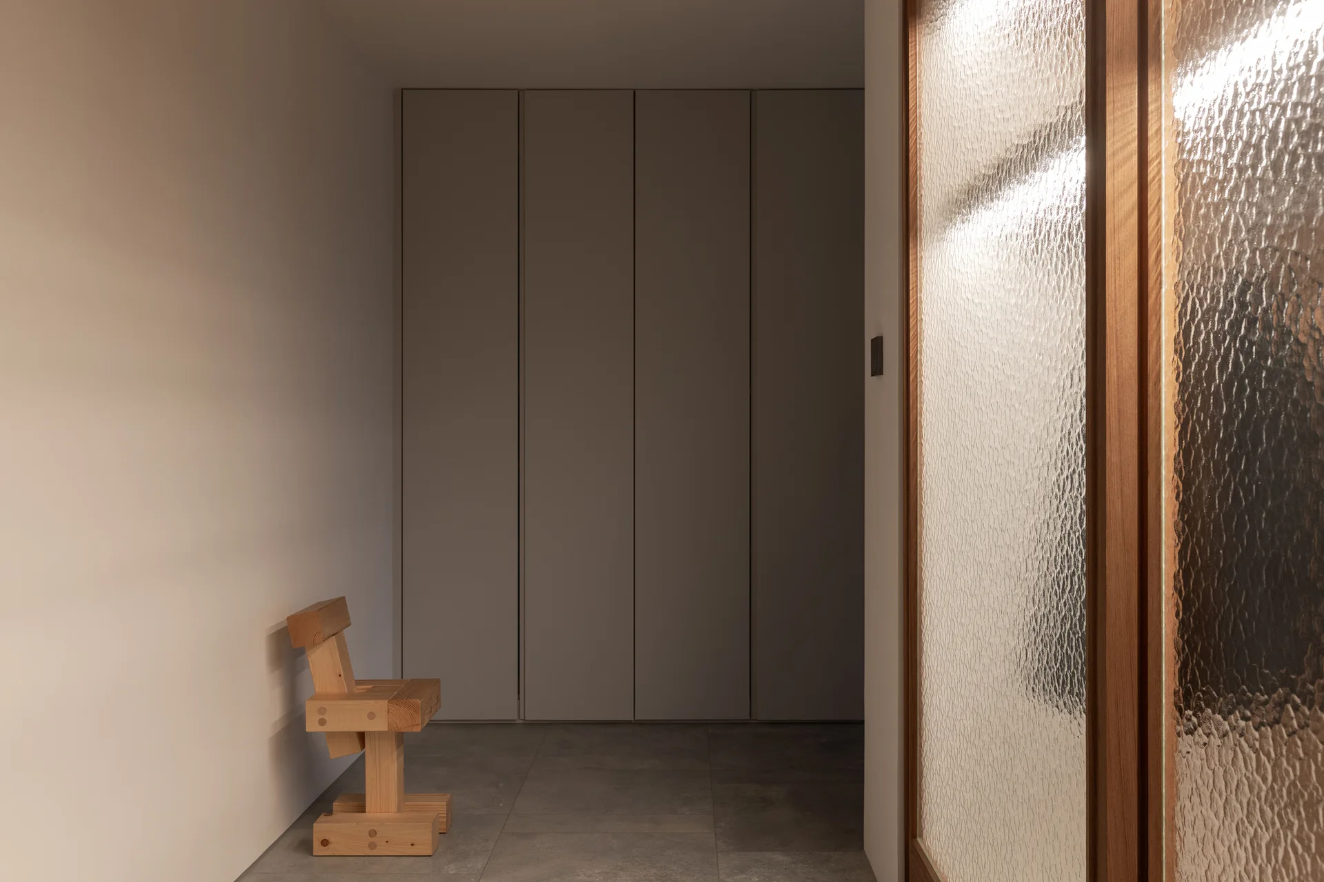 A minimalist hallway features large gray floor tiles, a sculptural wooden chair, full-height flush cabinets, and a textured glass partition.
