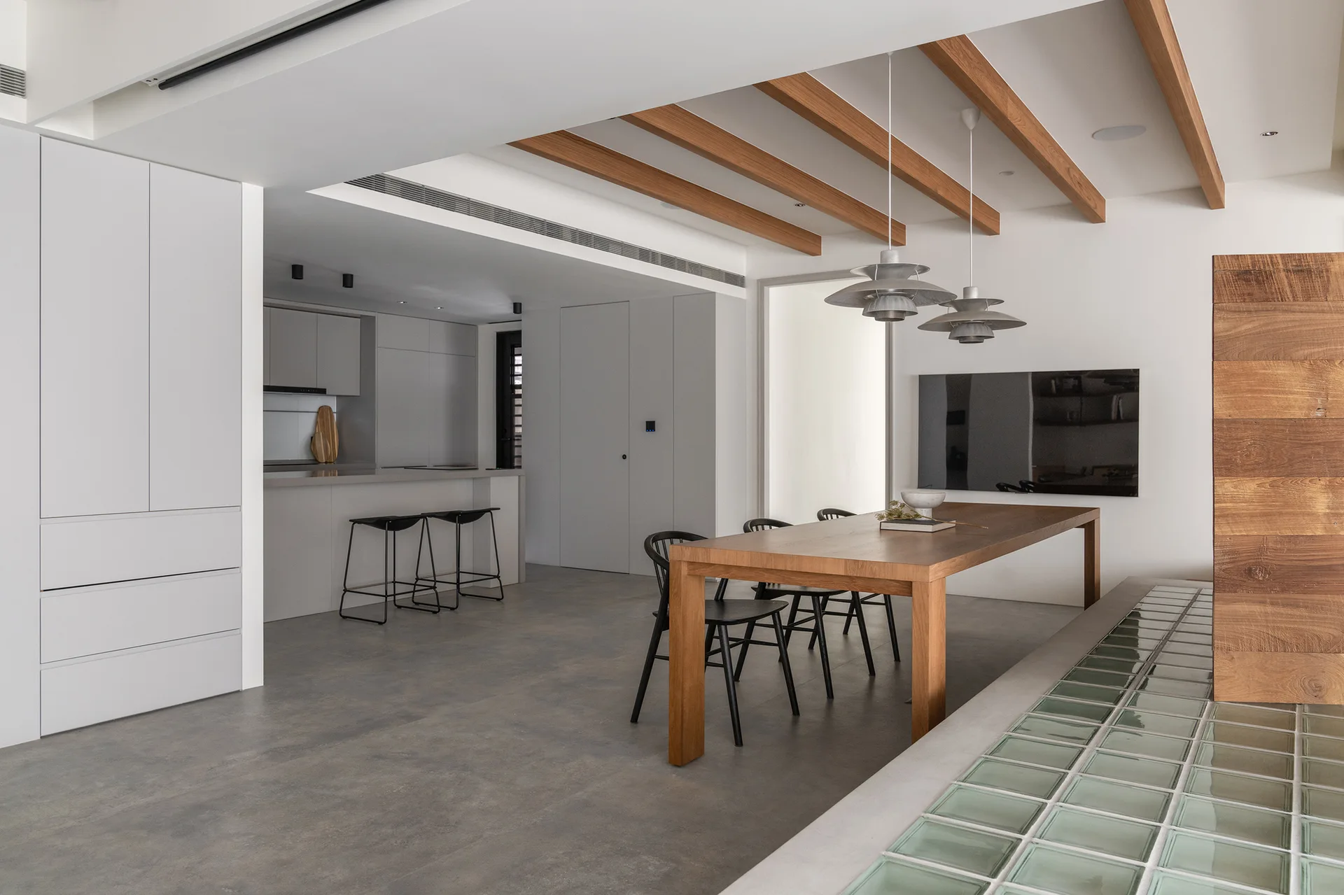 A minimalist dining area with a long wooden table, black chairs, concrete floors, and exposed wooden ceiling beams.