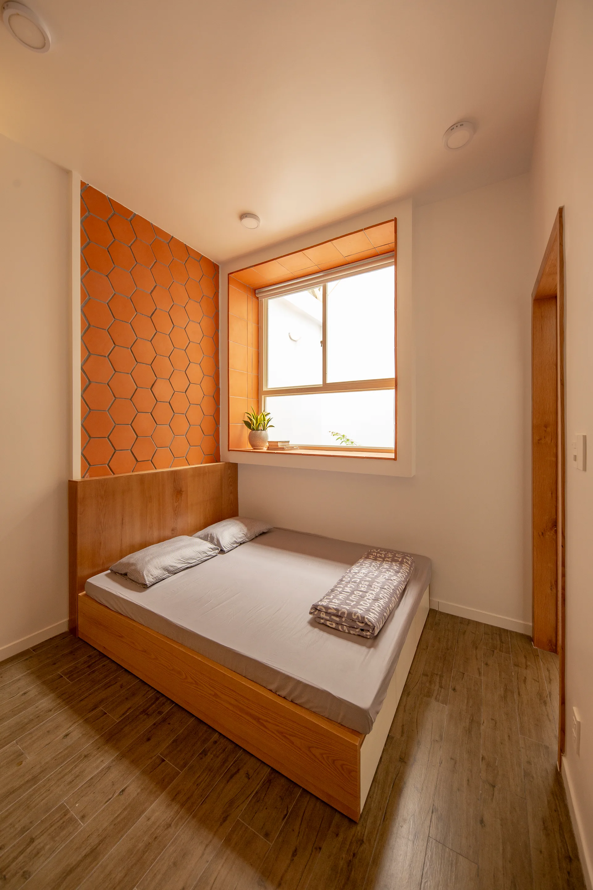 A wooden platform bed is positioned next to an orange hexagonal tile accent wall and a deep recessed window in a minimalist bedroom.