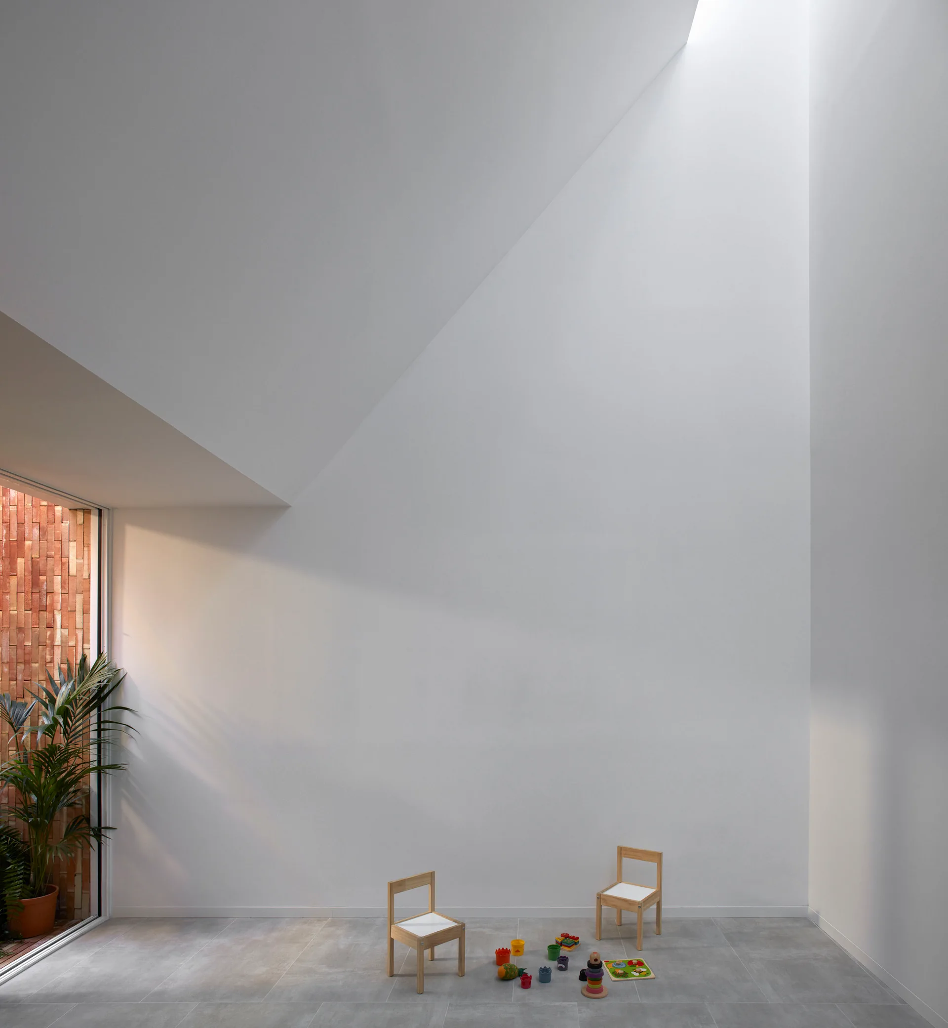 Two small wooden chairs and toys sit on grey tile floors in a minimalist white playroom with a high sloped ceiling and natural light.