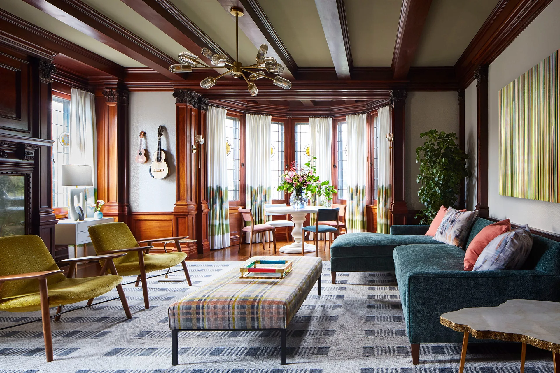 A living room features a teal sectional sofa, a plaid ottoman, mustard yellow armchairs, and dark wood wall paneling and ceiling beams.