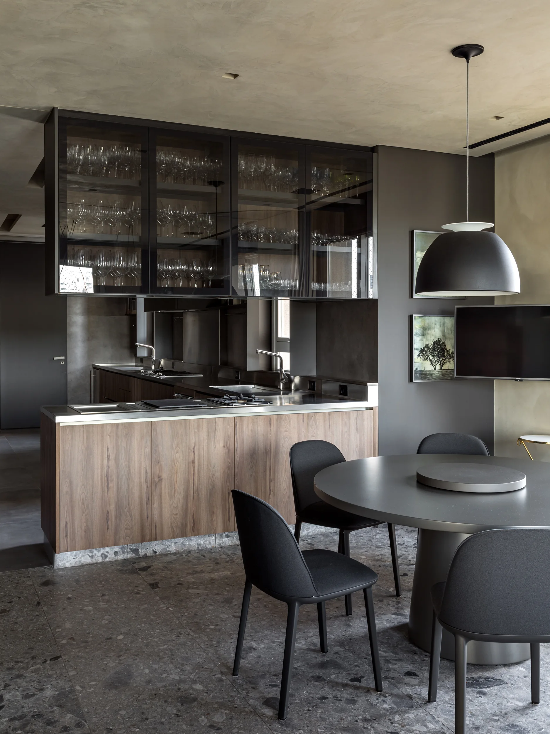 A modern charcoal kitchen features a wood-front island, suspended glass-front cabinets with glassware, and a round grey dining table.