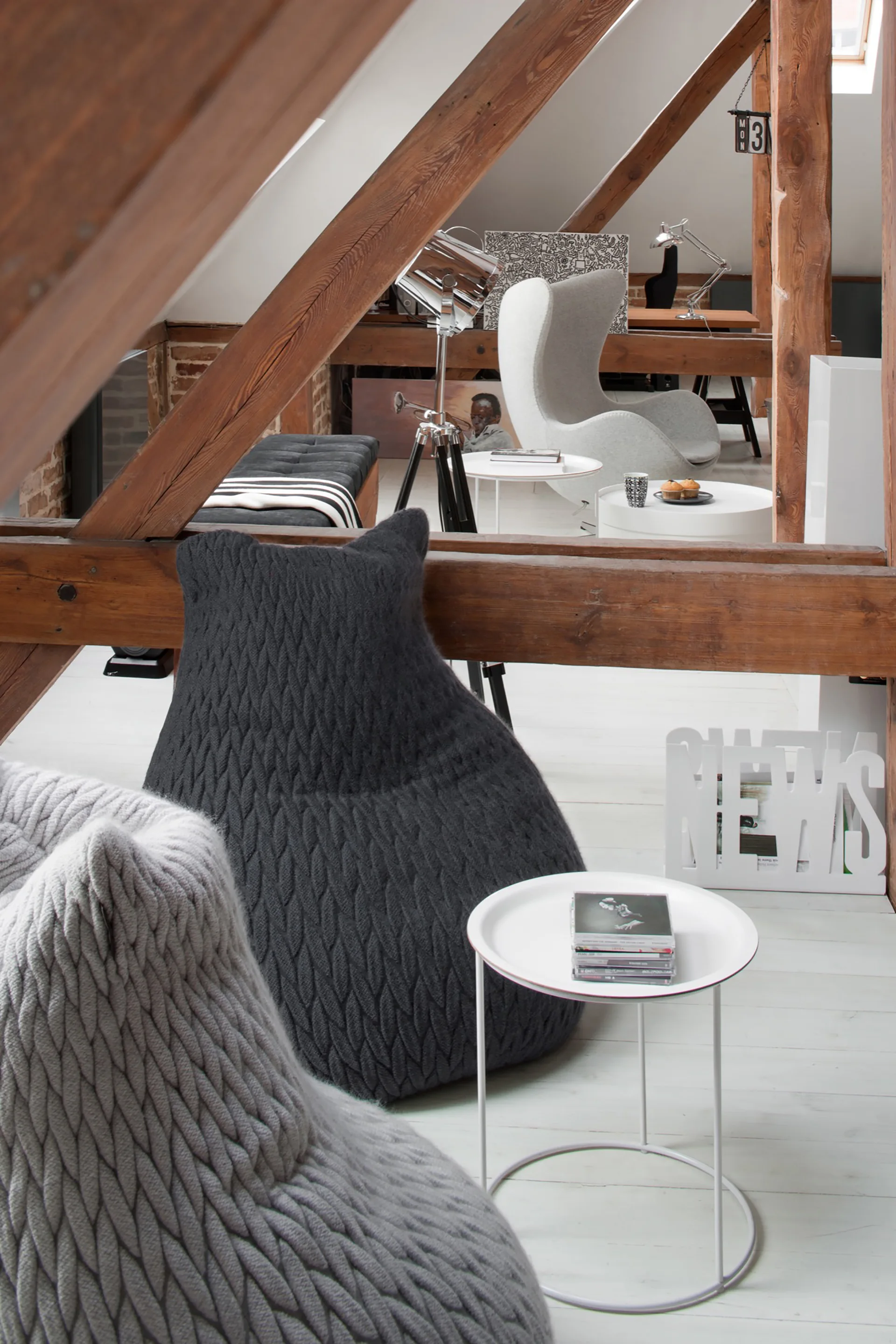 An attic loft space with dark wooden ceiling beams, white painted floorboards, and grey knitted bean bag chairs.