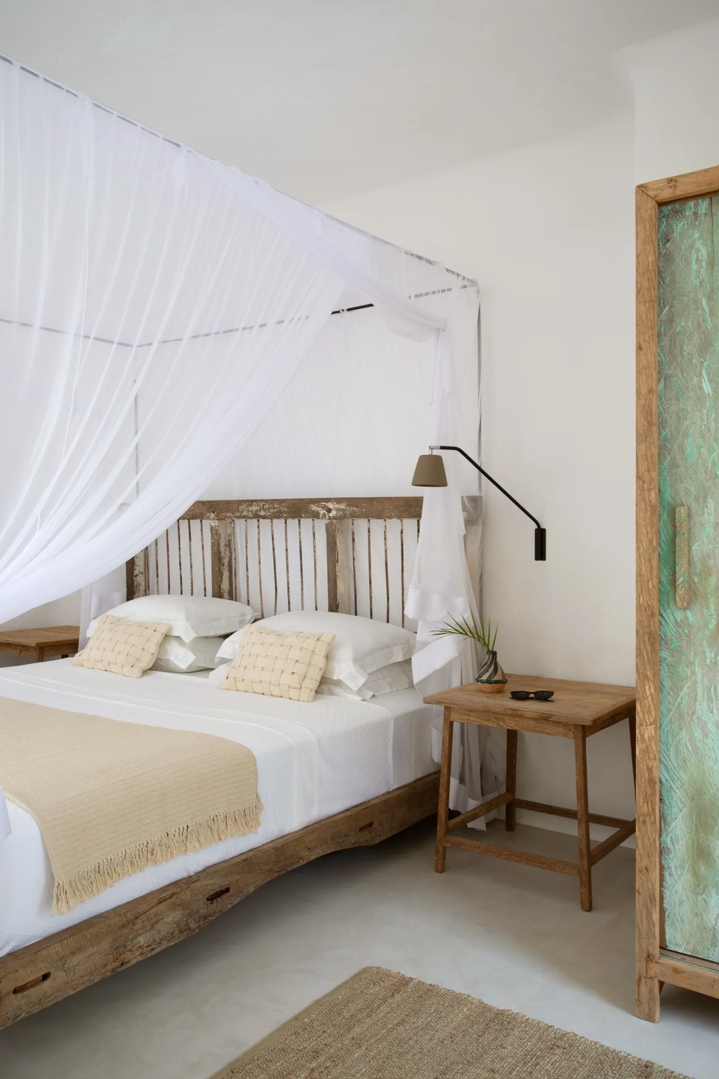 A rustic wooden canopy bed with sheer white draping sits next to a simple wooden nightstand under a black wall-mounted reading lamp.
