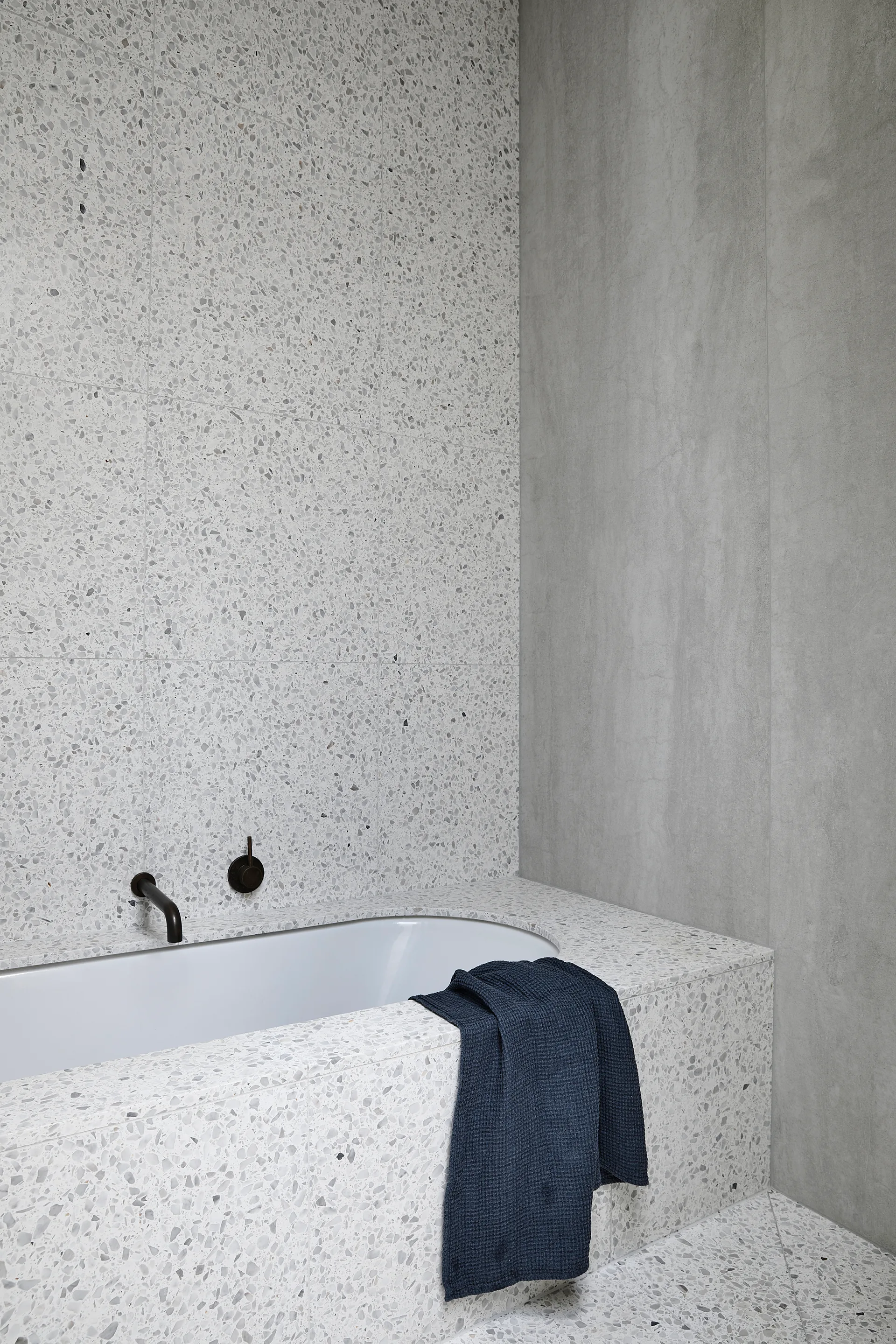 A built-in white bathtub is surrounded by speckled grey terrazzo tiles with a matte black faucet mounted on the wall and a blue towel on the edge.