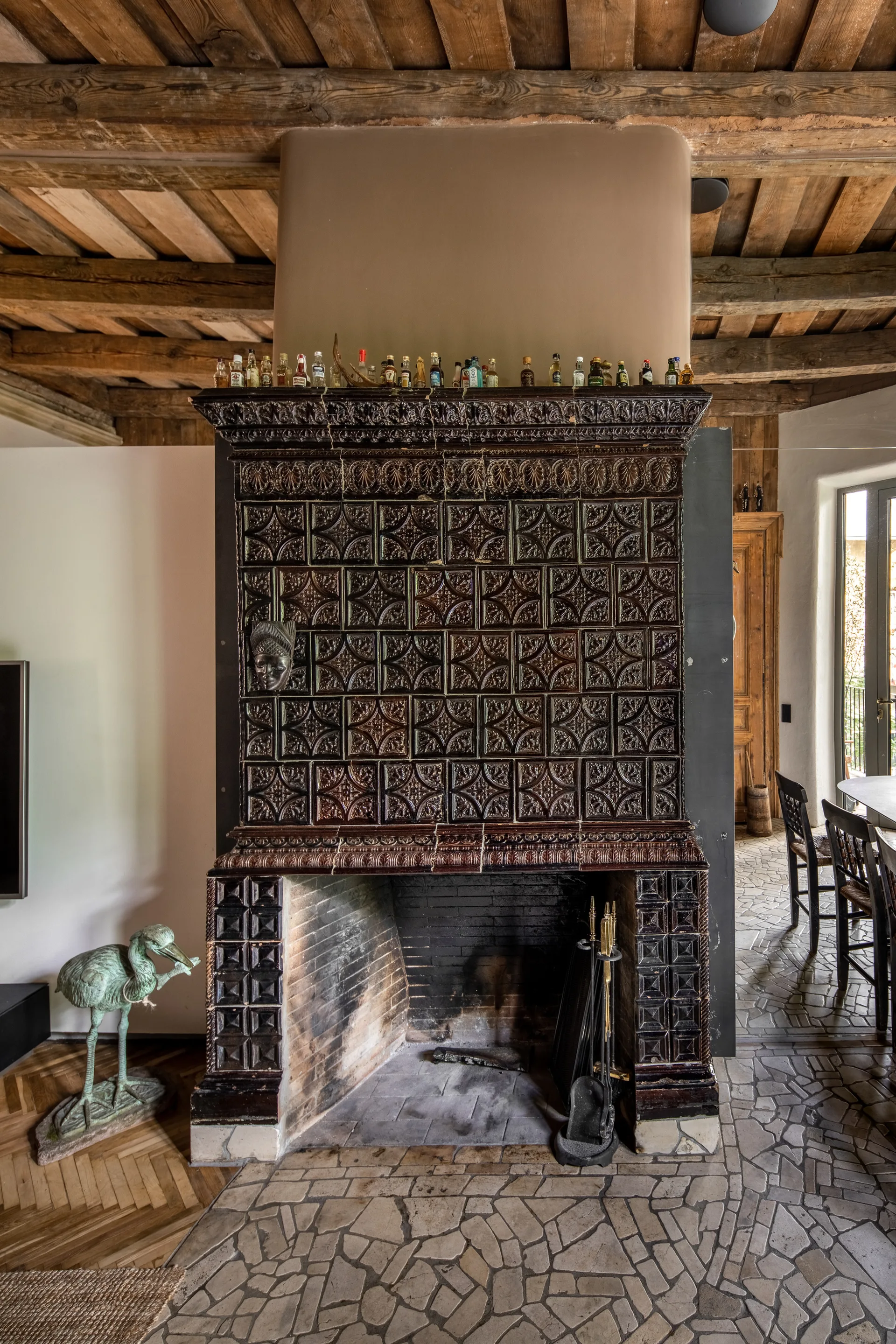 A large dark brown tiled fireplace with ornate relief patterns stands beneath an exposed wooden beamed ceiling on an irregular stone floor.