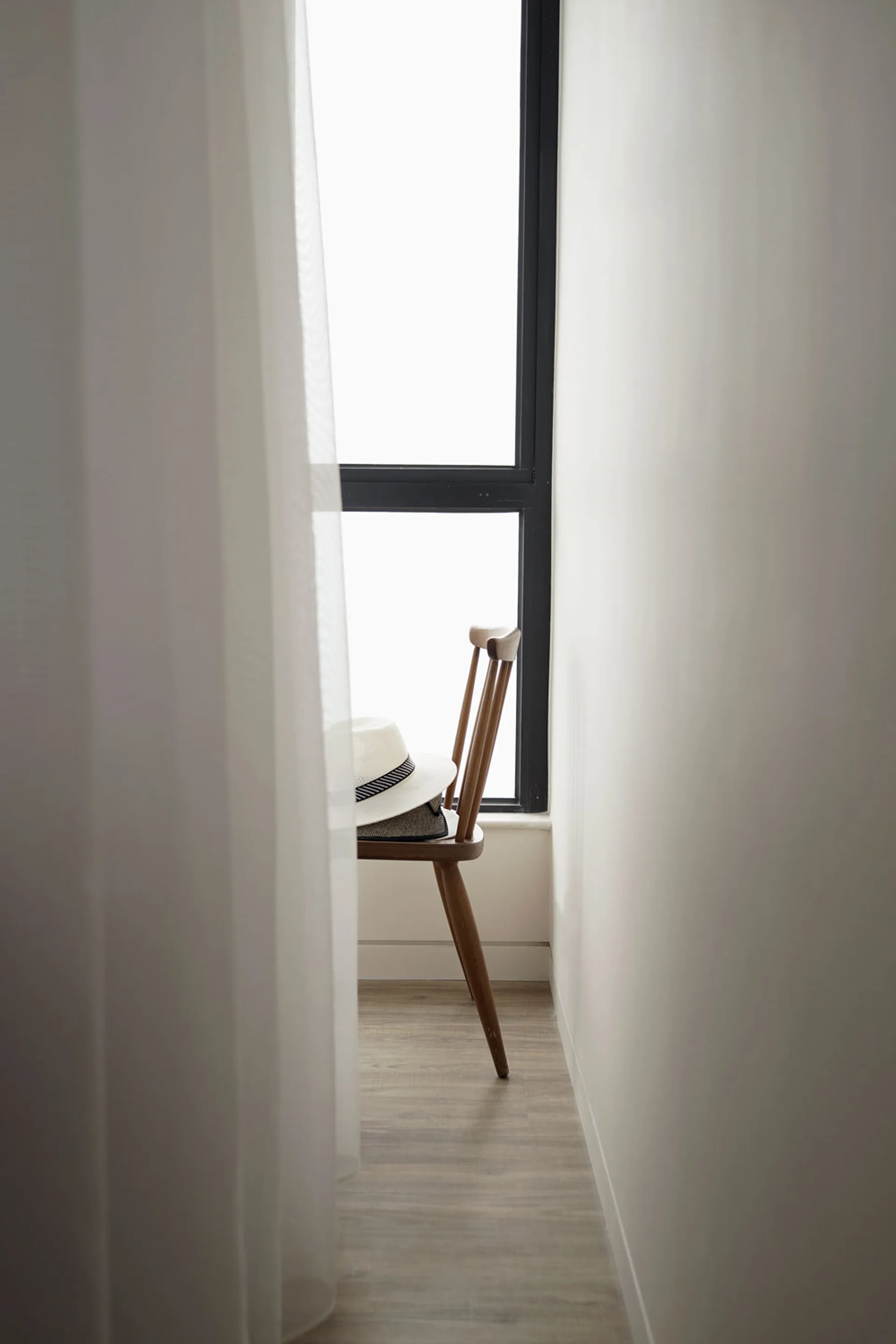 A mid-century modern wooden chair with a white straw hat sits in a narrow nook next to a tall black-framed window draped in sheer white curtains.