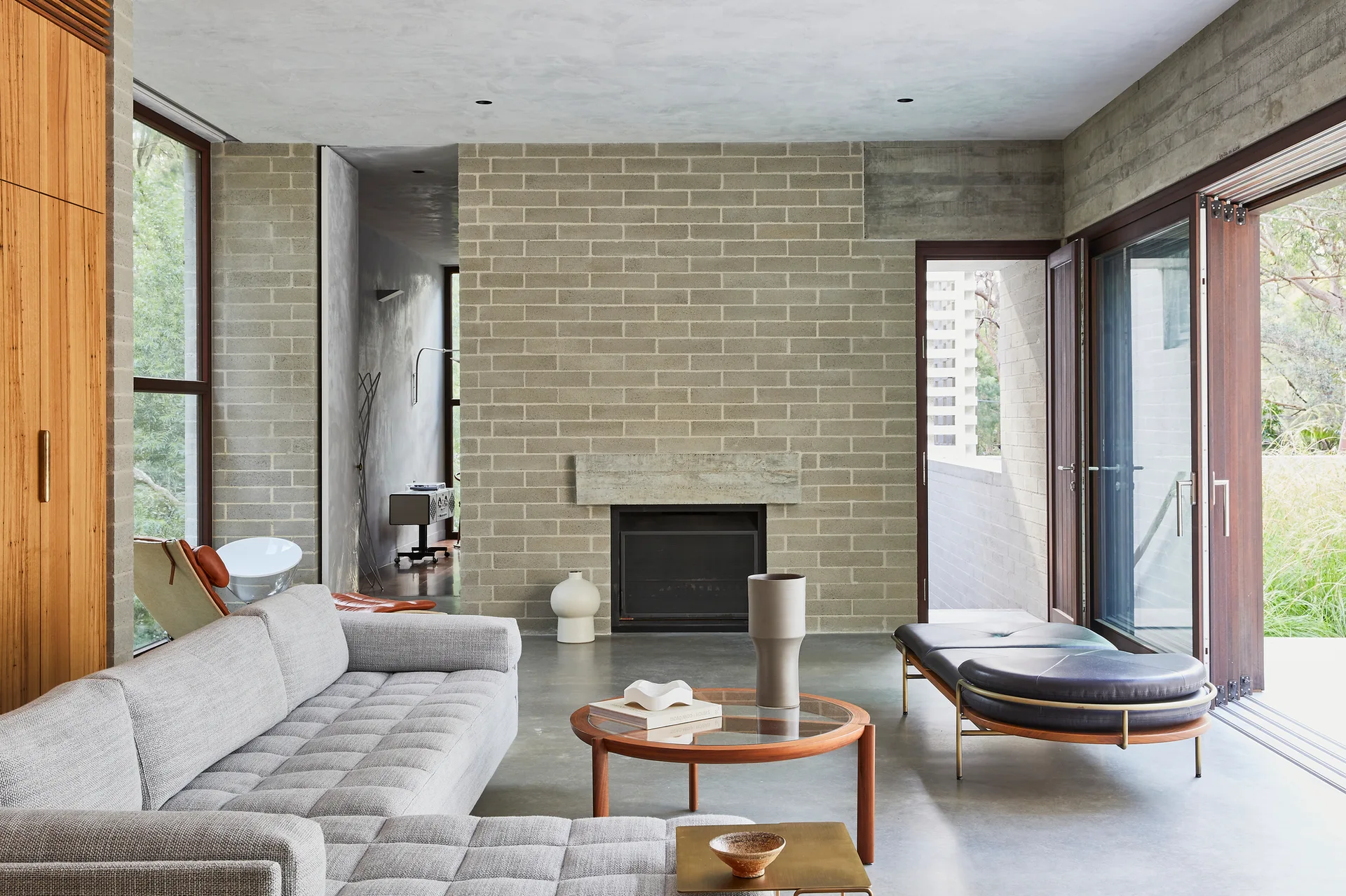 A modernist living room featuring a grey sectional sofa, a round wooden coffee table, and an exposed brick wall with a fireplace.