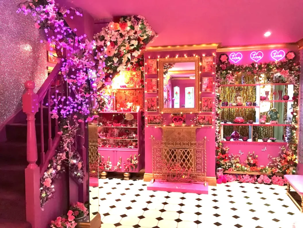 A bright pink living room featuring a black and white checkerboard floor, a pink staircase, a gold-framed mirror, and floral decorations.