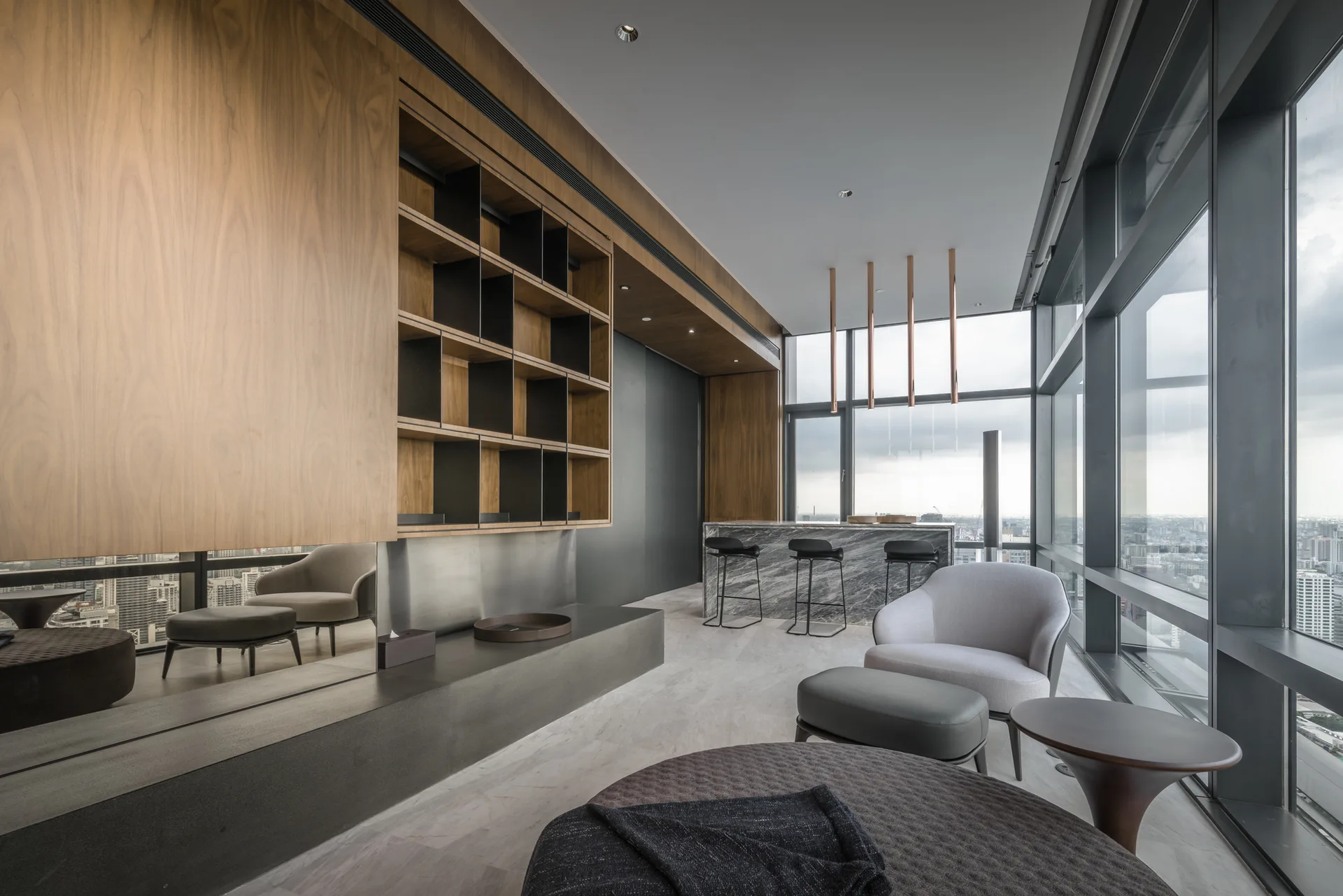 A modern high-rise lounge featuring custom wood wall shelving, a marble-patterned bar counter with stools, and floor-to-ceiling windows.