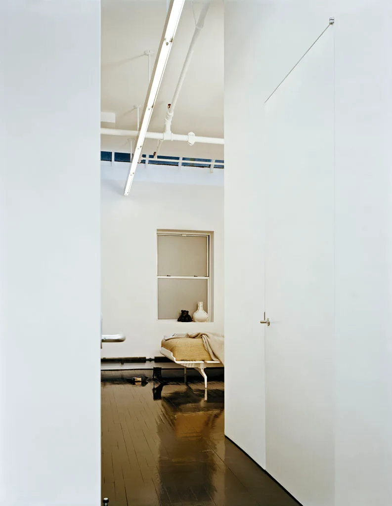 A view looking into a minimalist white bedroom featuring a low-profile bed, dark glossy wood flooring, and exposed white pipes on the ceiling.