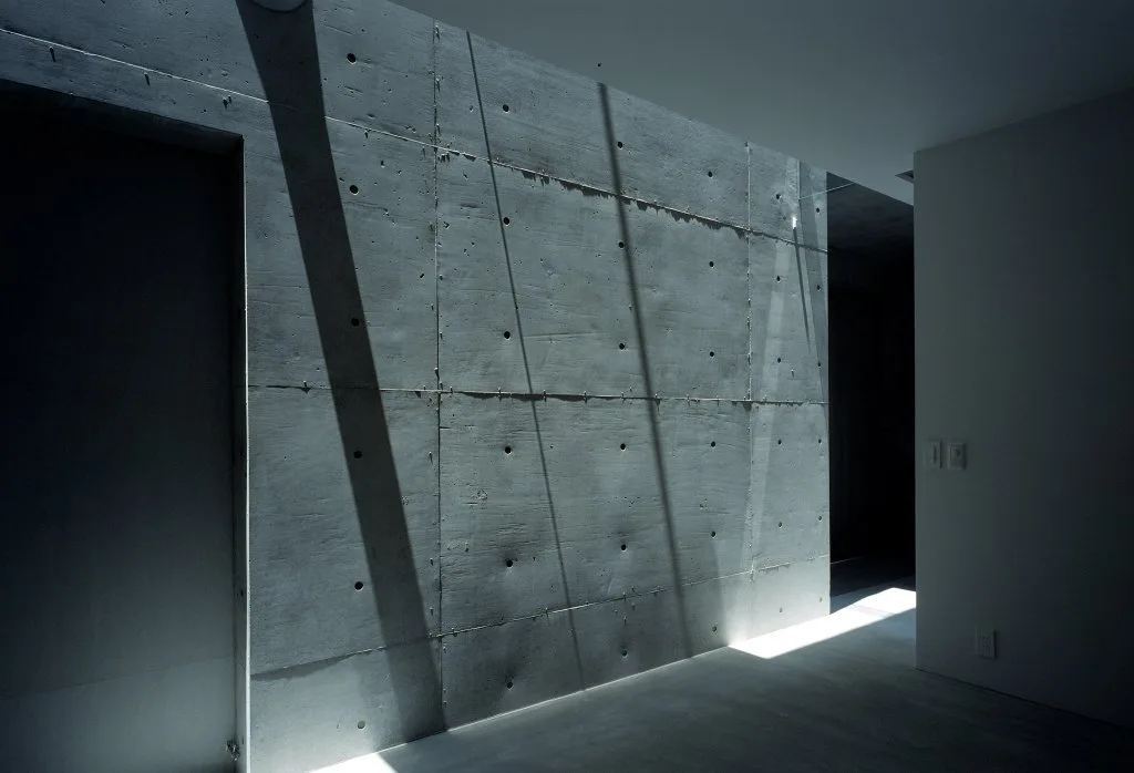 A minimalist hallway features a raw concrete wall with a grid of formwork holes under dramatic natural sunlight and shadows.