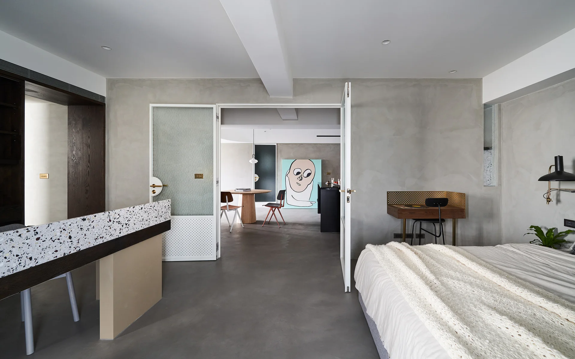 A modern bedroom with gray concrete walls features a bed with white knit throw, a terrazzo console, and open glass doors leading to a dining area.
