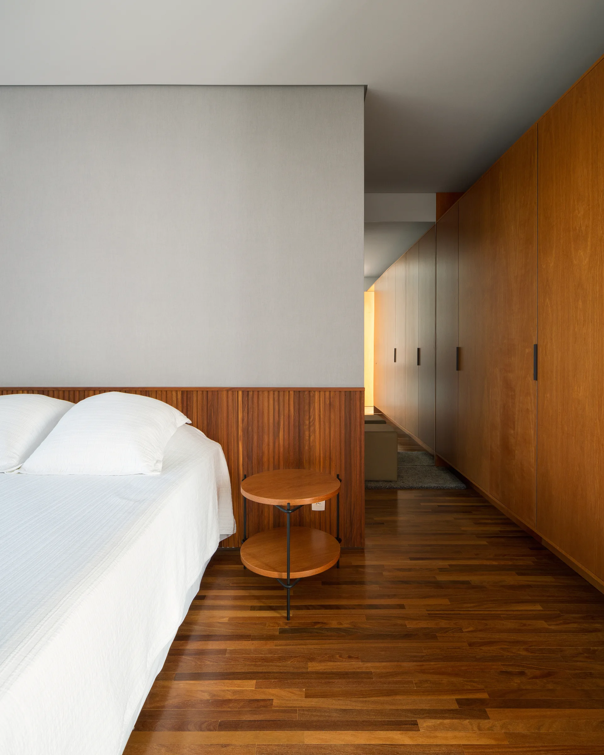 A minimalist bedroom features a white bed with a wooden slatted headboard panel, a round two-tier side table, and a built-in wooden wardrobe wall.