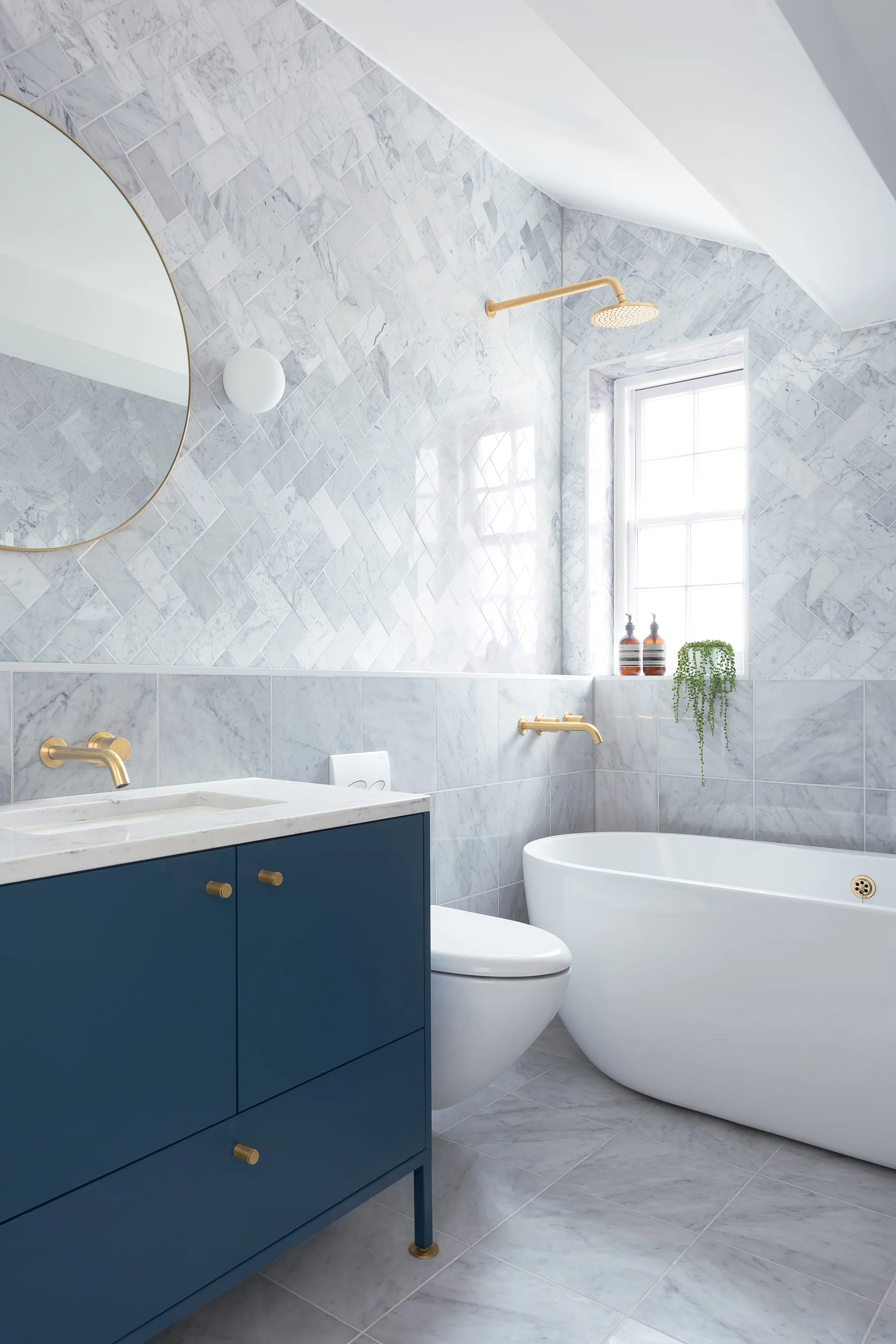 A modern bathroom featuring a navy blue vanity, a freestanding white bathtub, marble herringbone wall tiles, and brushed gold fixtures.