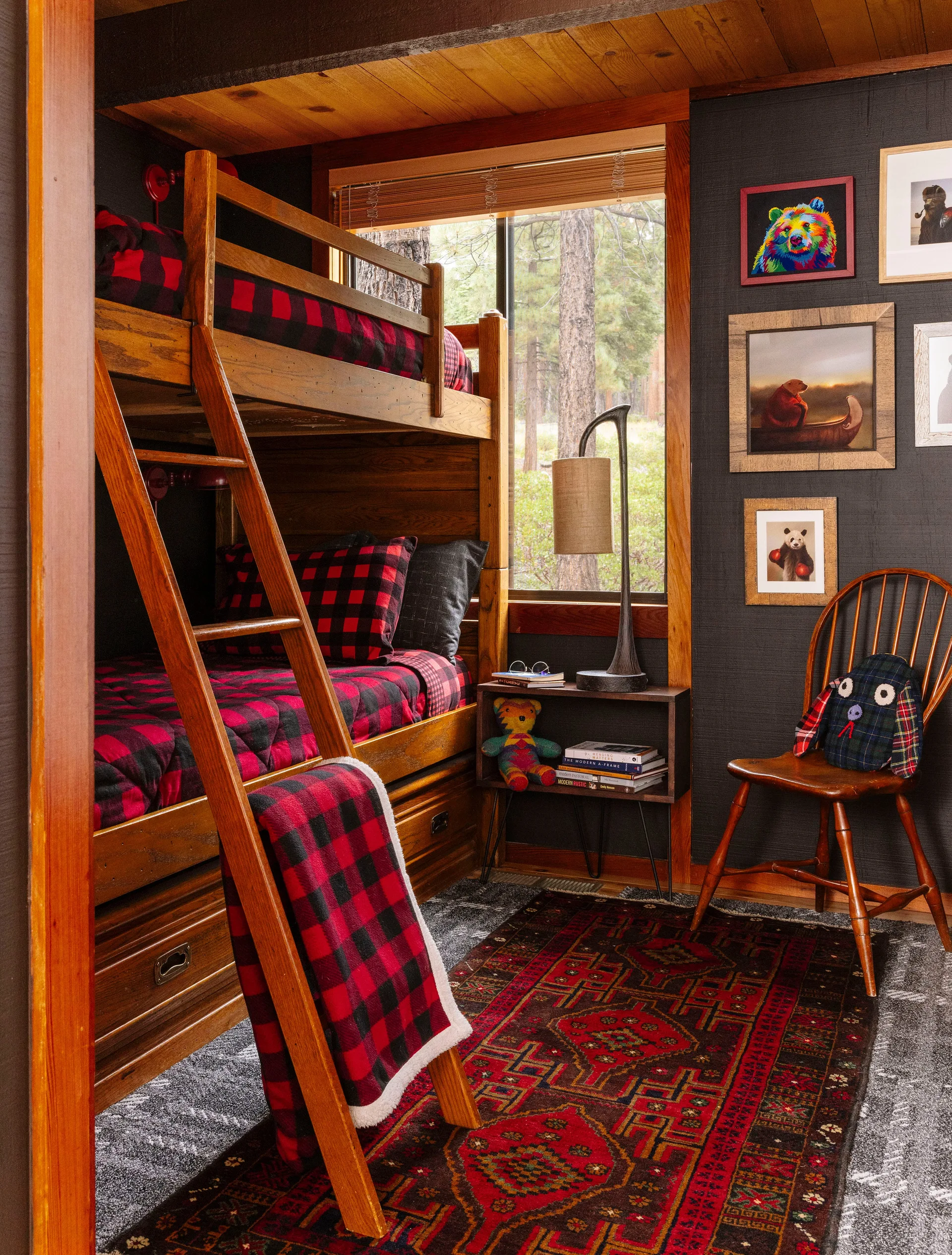 A rustic bedroom features a wooden bunk bed with red and black buffalo check bedding next to a dark wall and window.