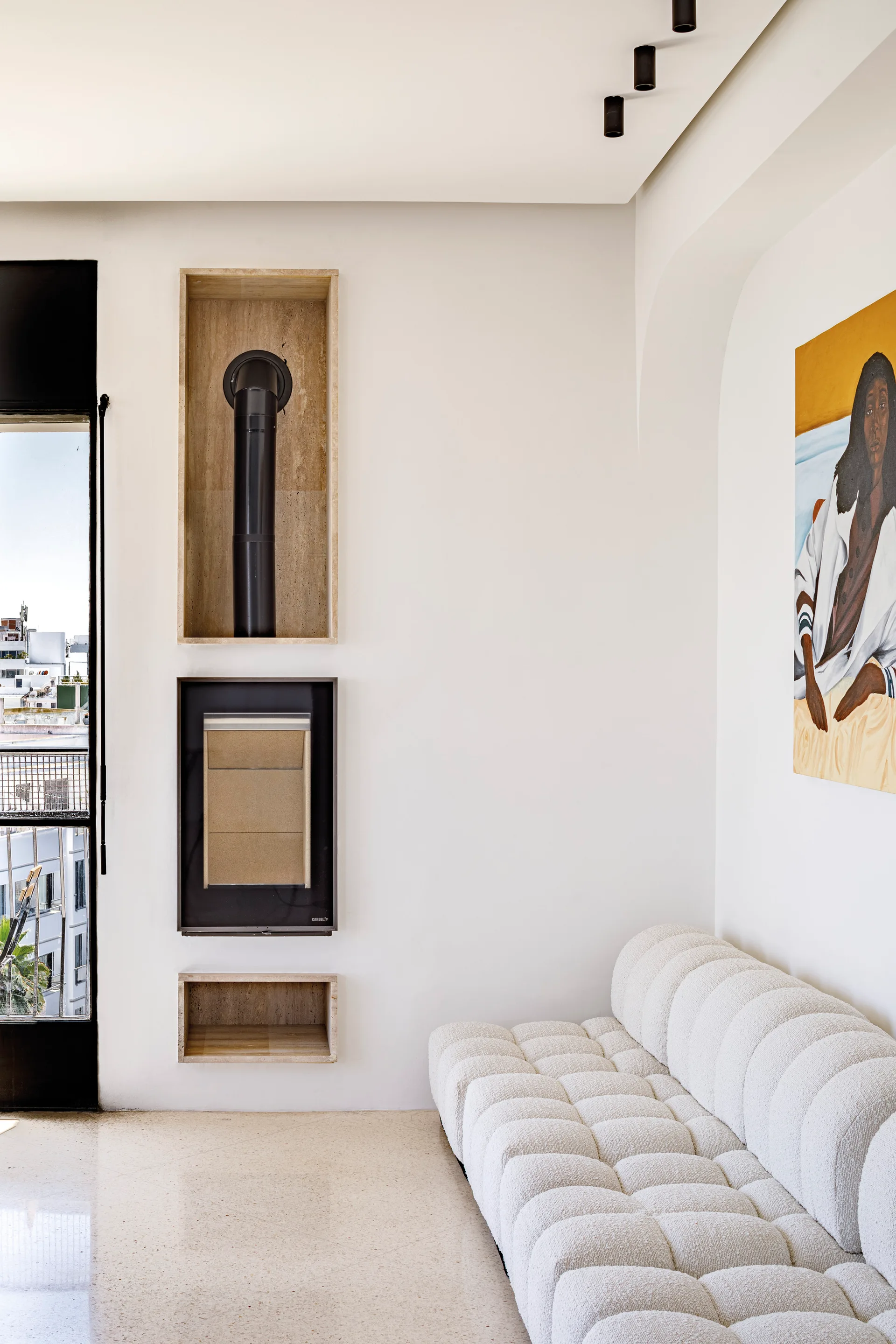A minimalist white room features a tufted cream sofa alongside an integrated vertical fireplace set into stone-lined wall niches.