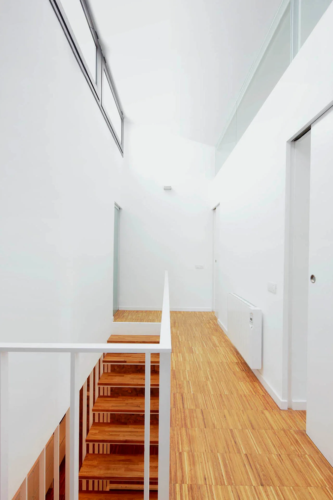 A minimalist white hallway featuring wooden floors and a matching wooden staircase with a simple white metal railing.