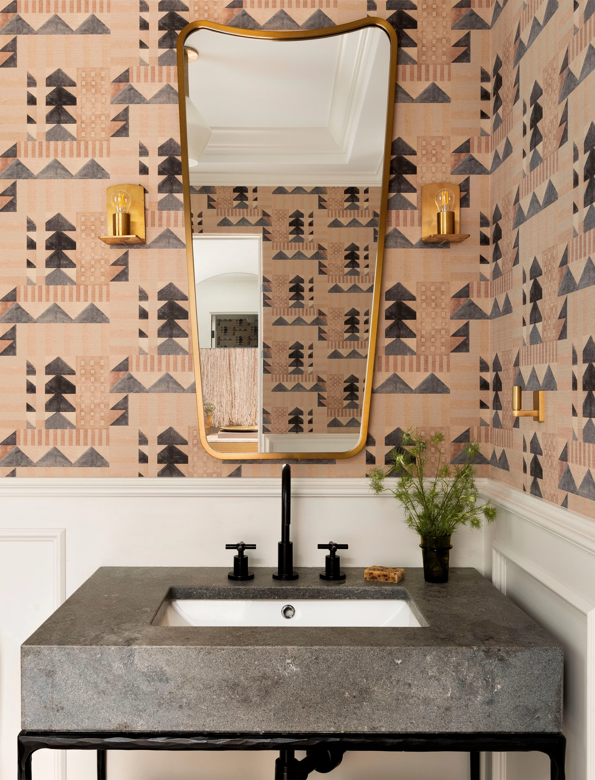 A powder room features tan geometric wallpaper above white wainscoting, a textured grey stone console vanity, and a brass-framed mirror flanked by sconces.