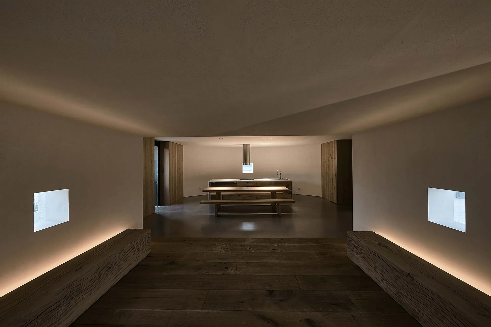 A minimalist kitchen and dining area features a wooden island and matching dining table flanked by low benches with integrated cove lighting.